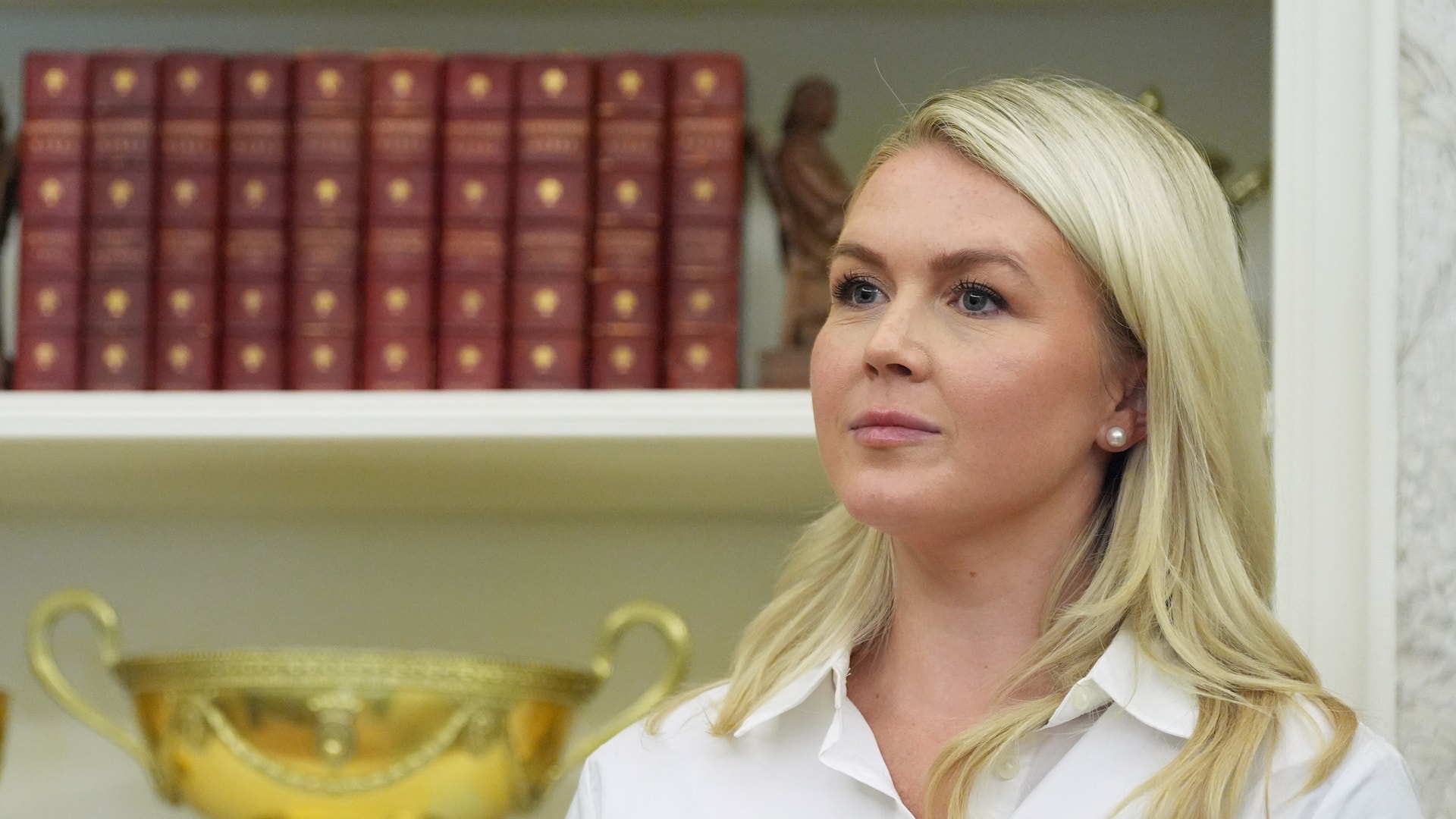 Porträtt av en kvinna med blont hår i vit skjorta framför en bokhylla med röda böcker och en gyllene skål.