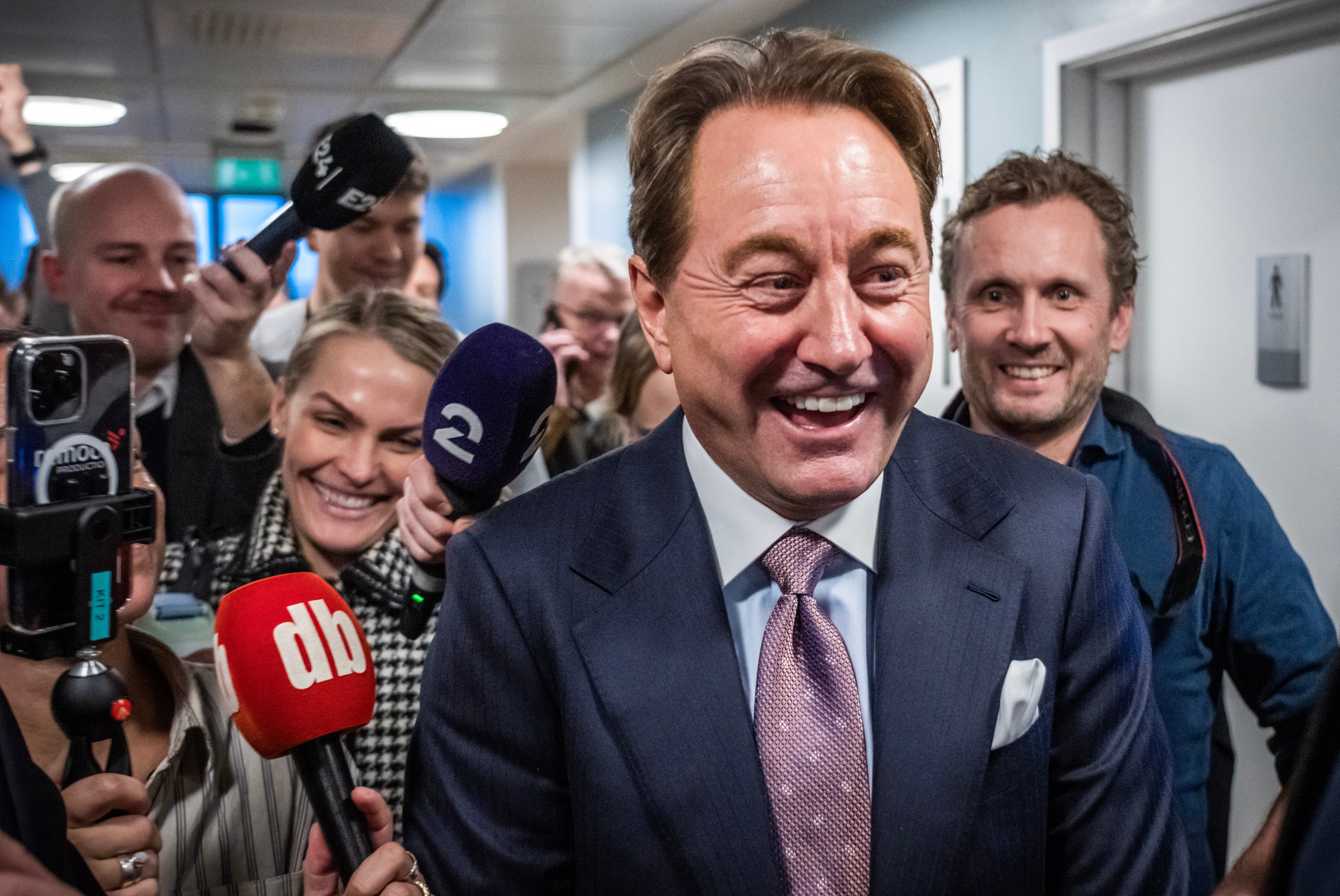 En leende man i kostym omringad av journalister med mikrofoner i en korridor.