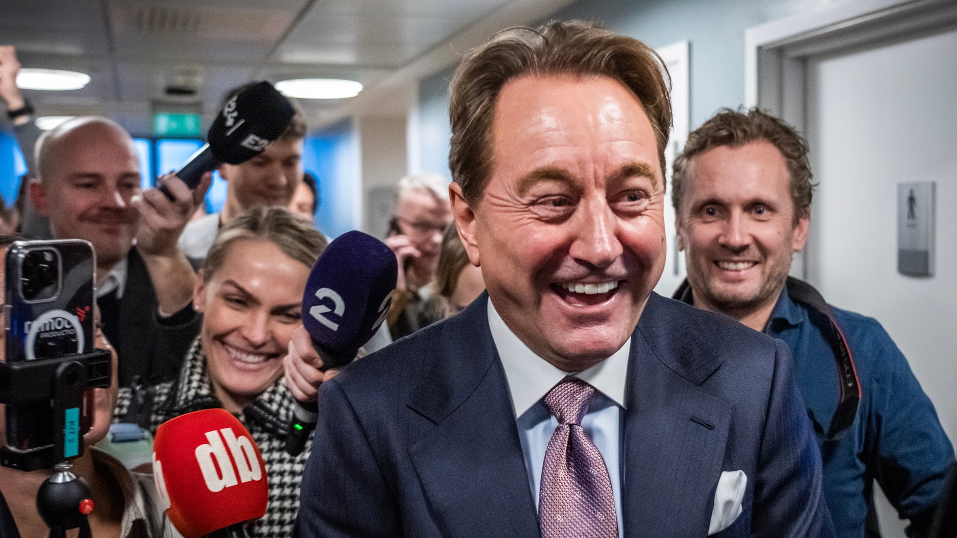 En leende man i kostym omringad av journalister med mikrofoner i en korridor.