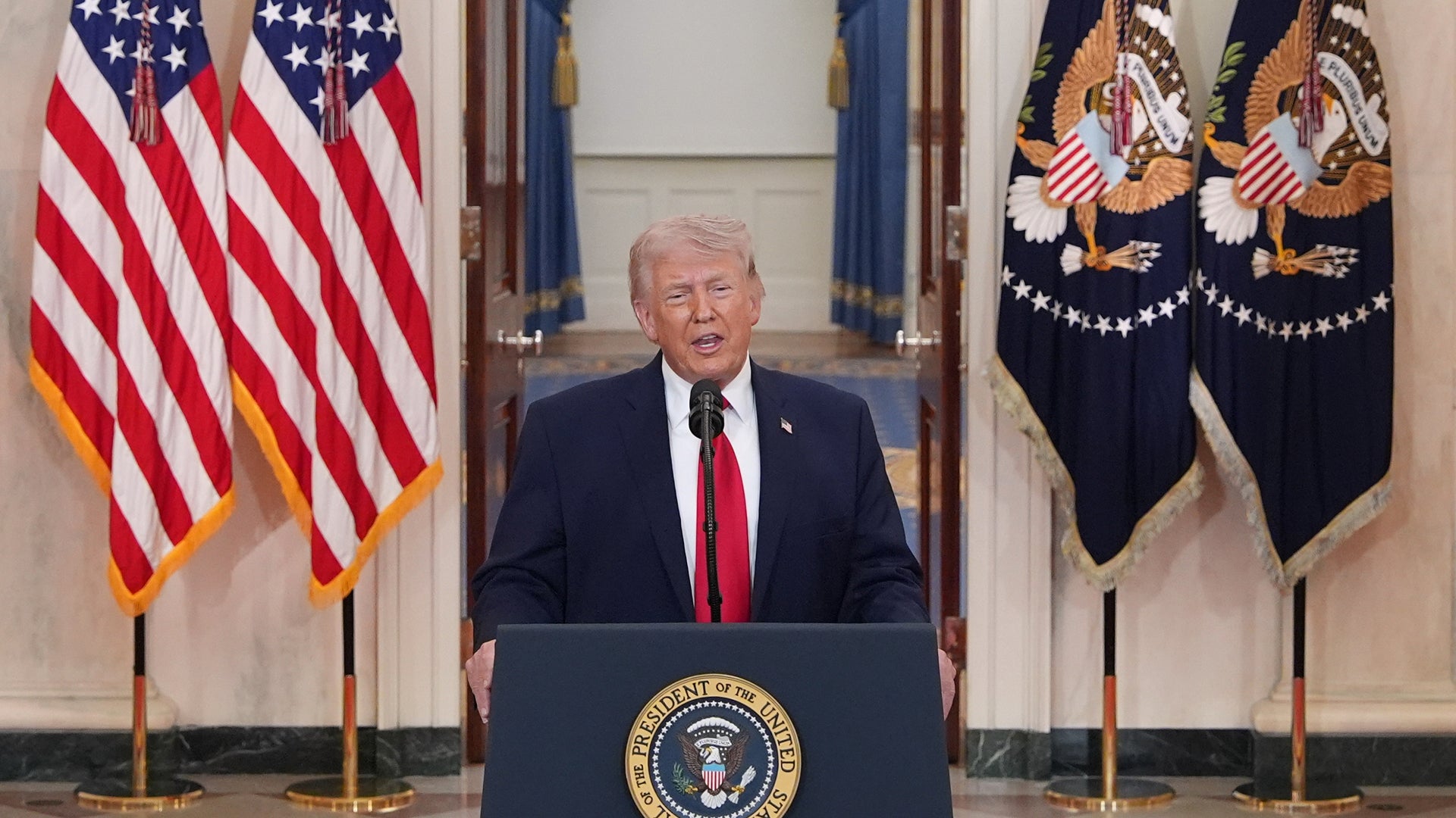 Man i mörk kostym och röd slips står vid ett presidentpodium omgivet av amerikanska flaggor.