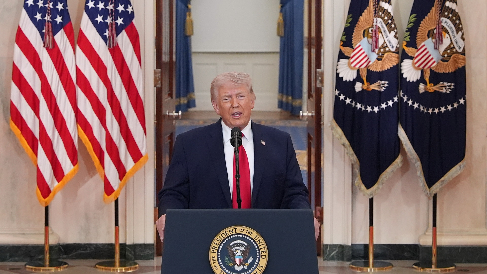 Man i mörk kostym och röd slips står vid ett presidentpodium omgivet av amerikanska flaggor.