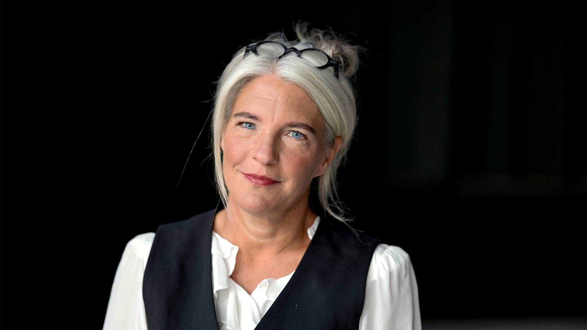 Porträtt av en leende kvinna med gråblont hår mot en mörk bakgrund.
