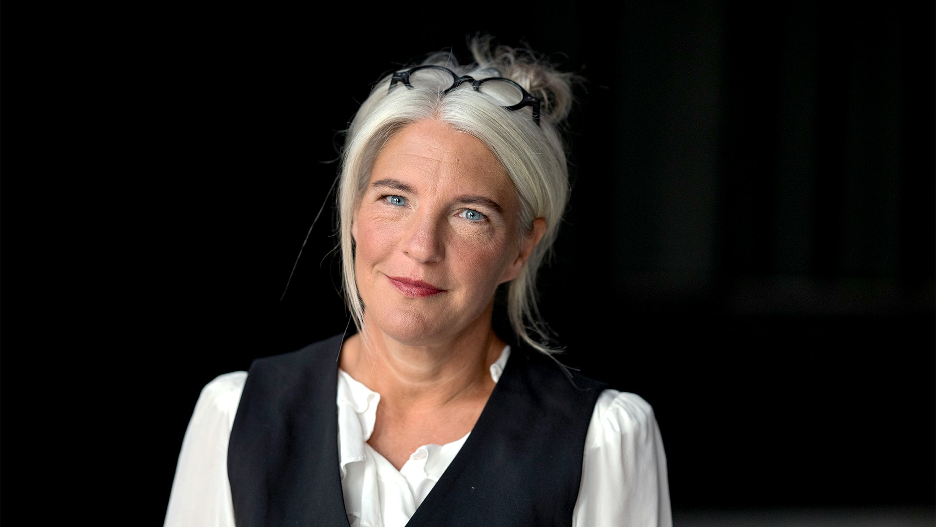 Porträtt av en leende kvinna med gråblont hår mot en mörk bakgrund.