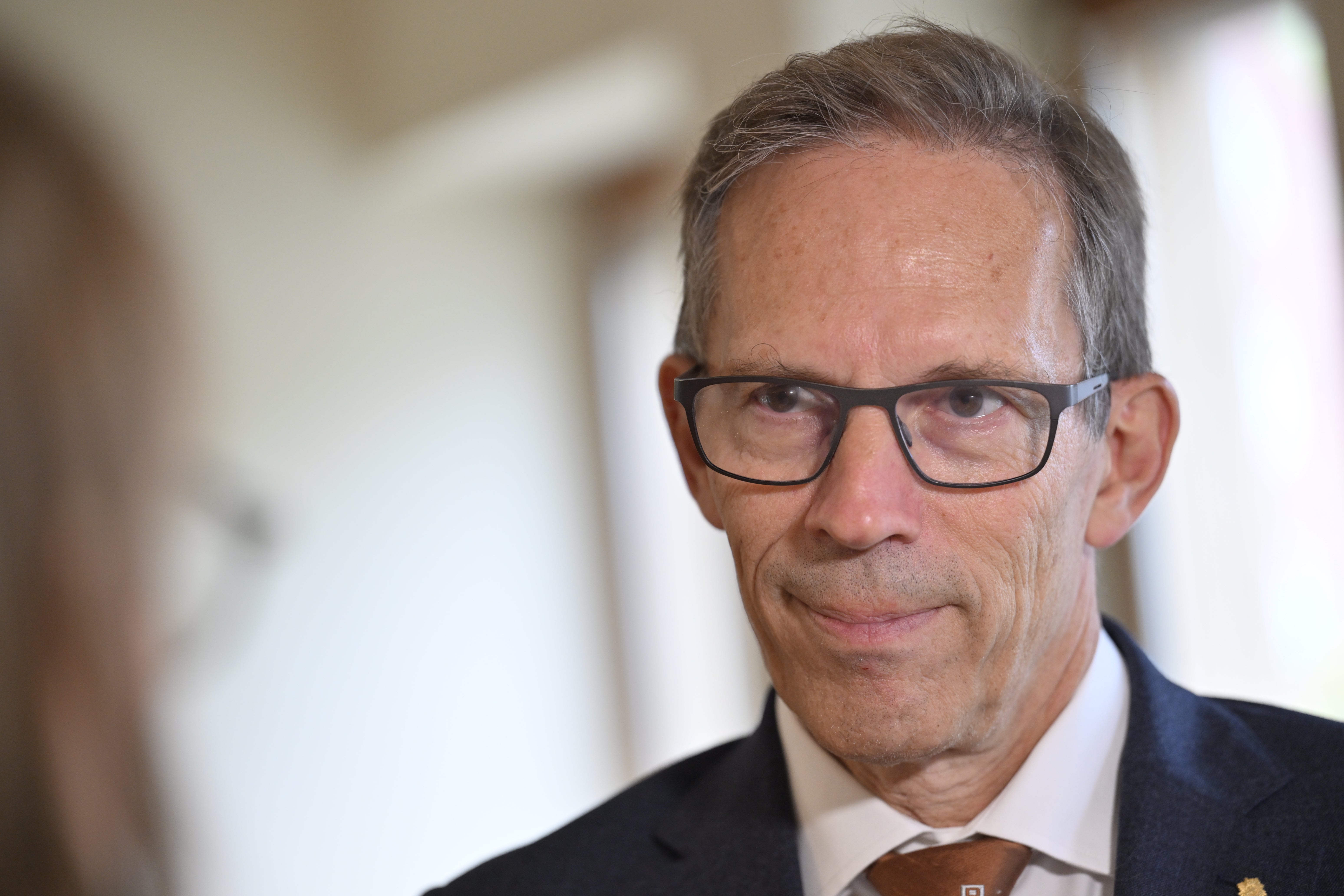 En äldre man i kostym och glasögon tittar mot kameran under ett samtal med en person i förgrunden.