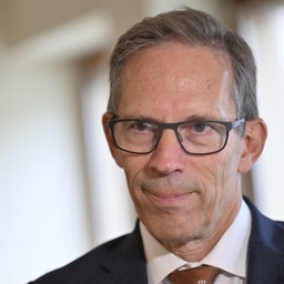 En äldre man i kostym och glasögon tittar mot kameran under ett samtal med en person i förgrunden.