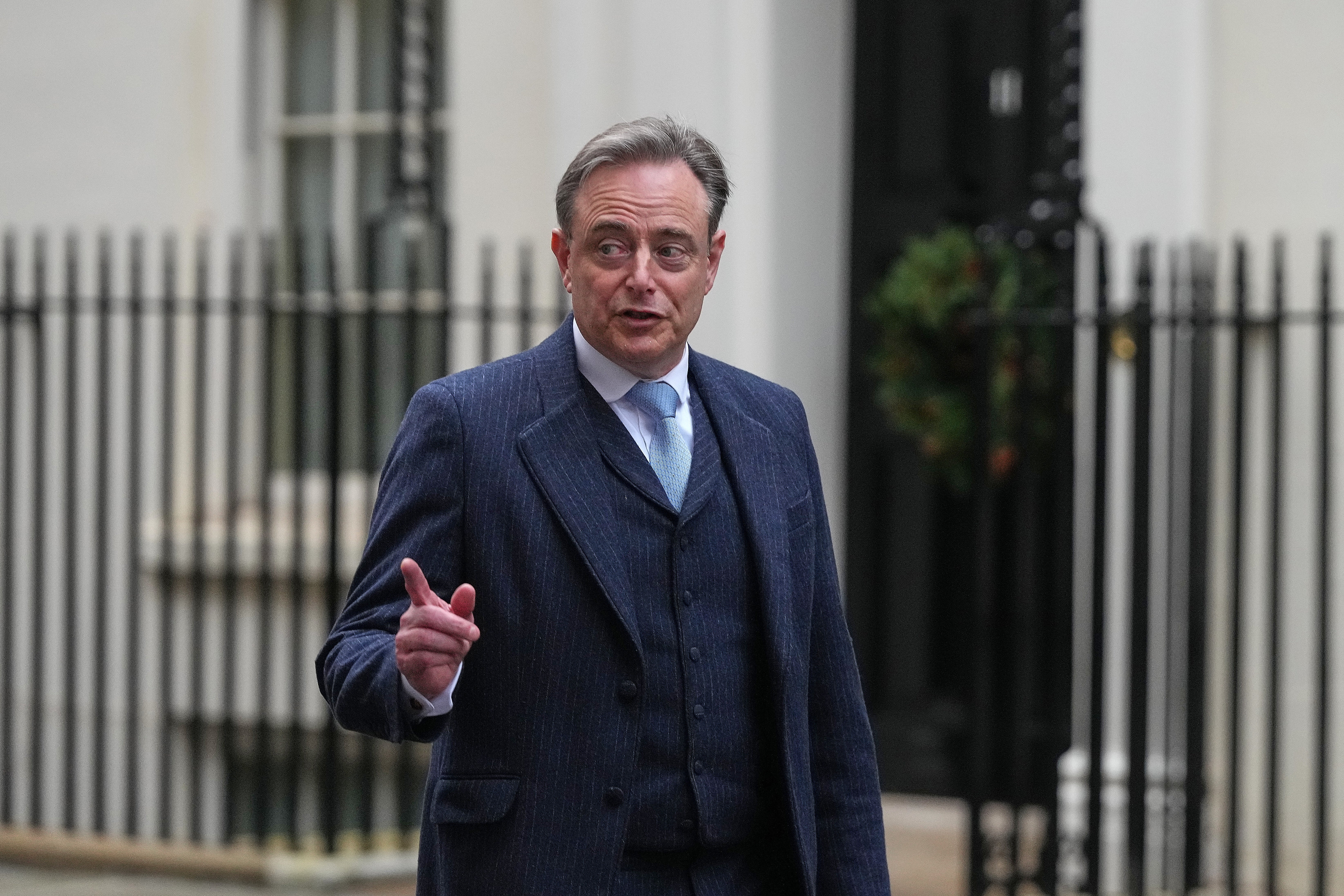 En man i mörkblå kostym och glasögon framför en vit byggnad med järnstaket.