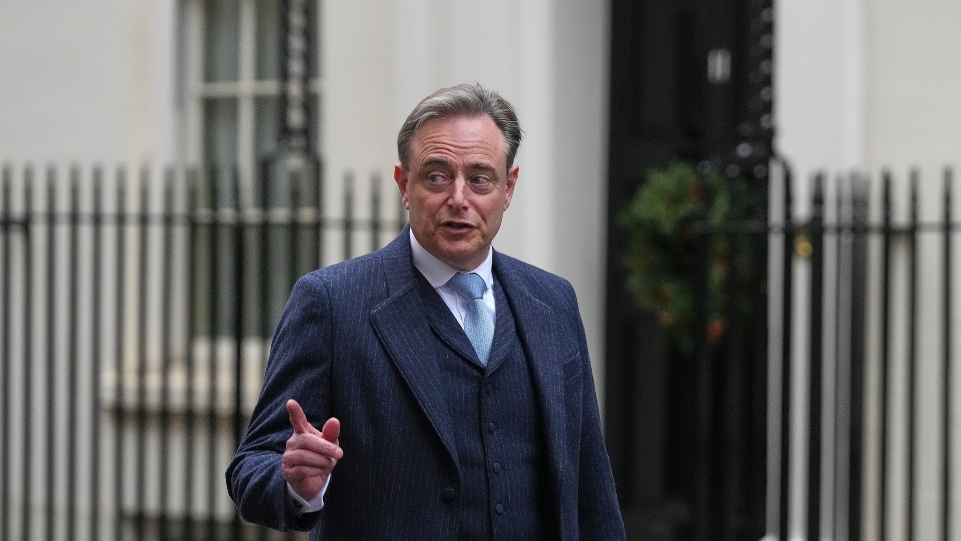 En man i mörkblå kostym och glasögon framför en vit byggnad med järnstaket.