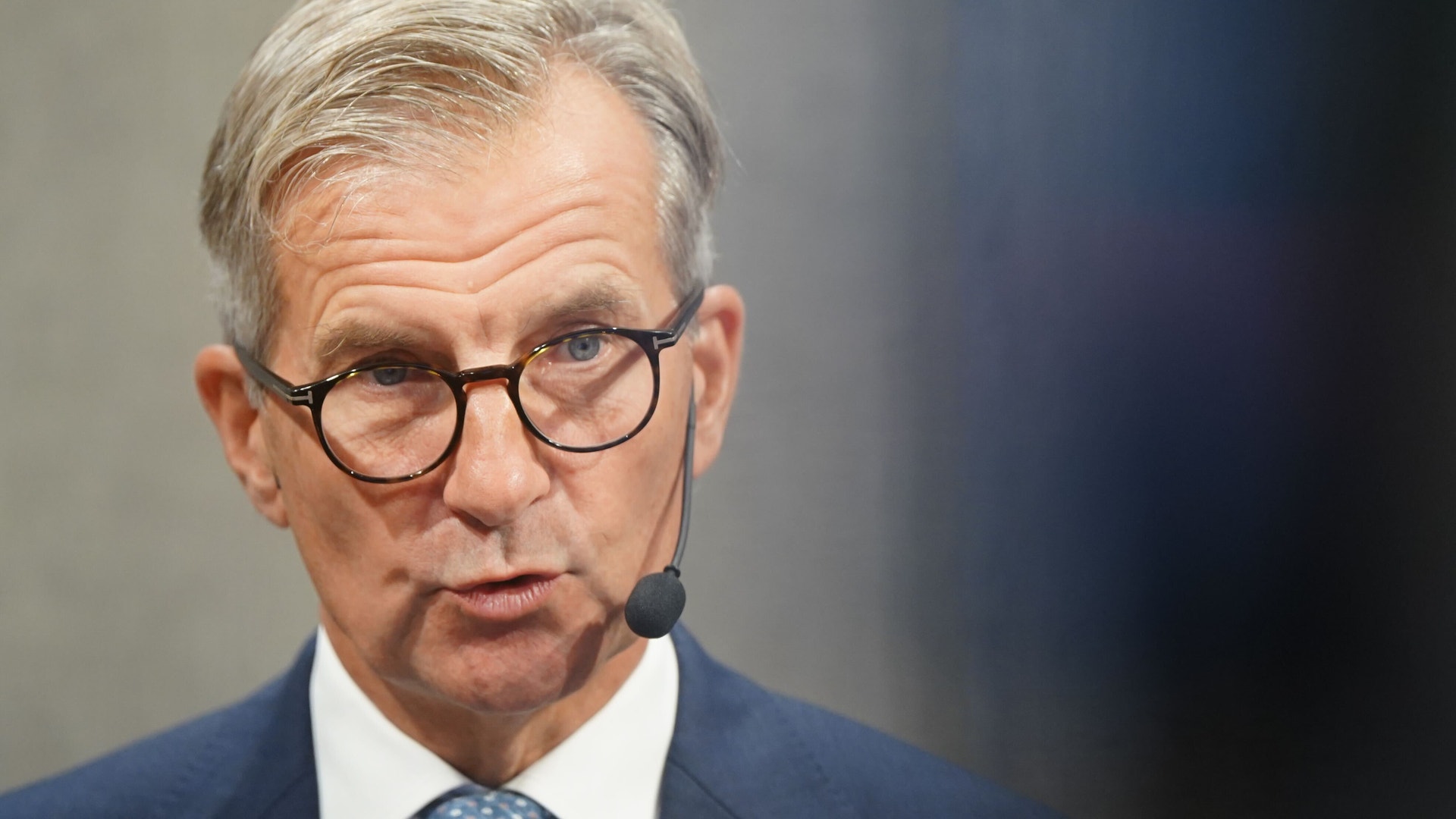 En man i glasögon och mörkblå kostym talar vid en mikrofon