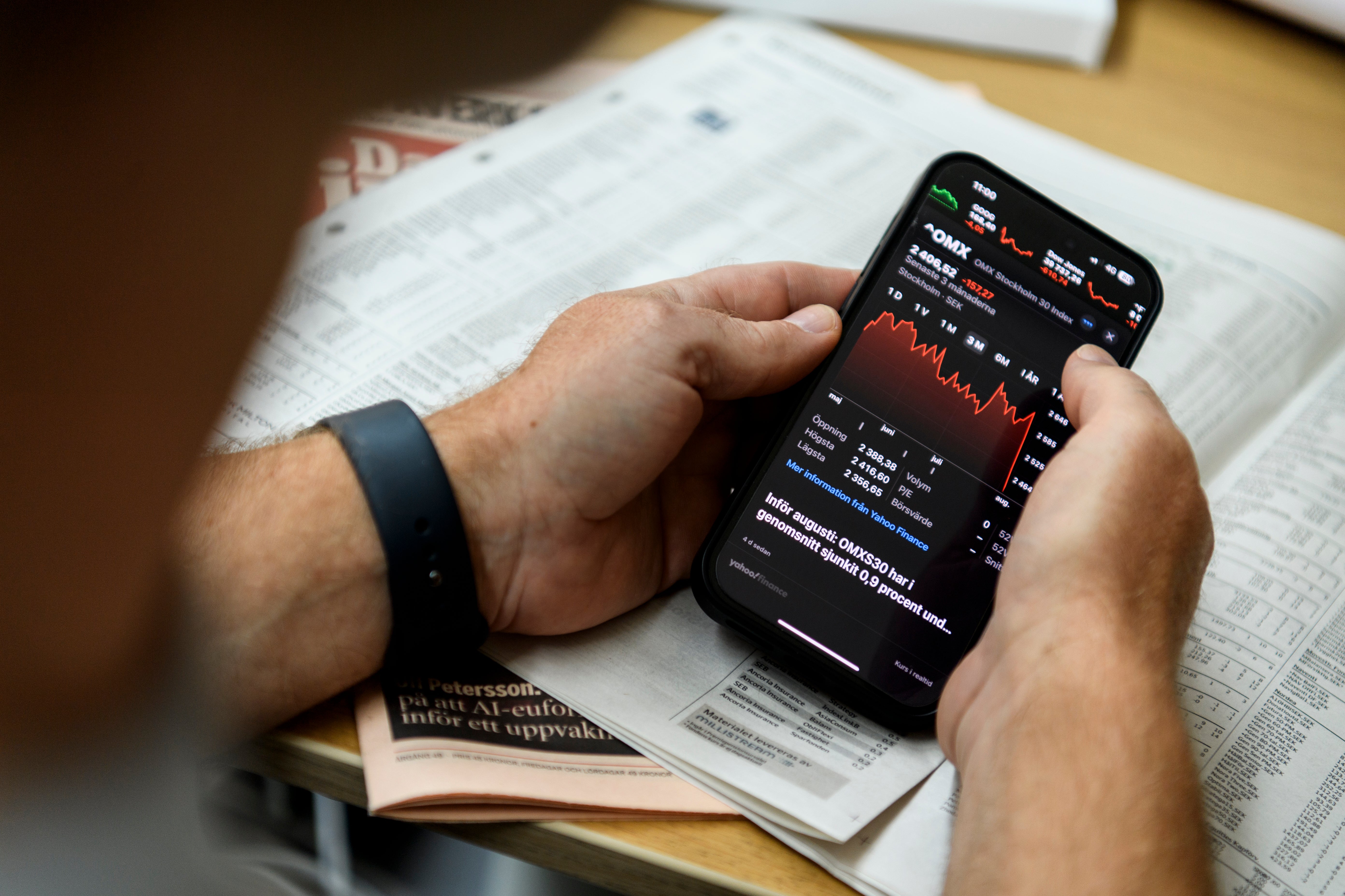 Person håller en smartphone med aktiemarknadsinformation och grafer, sittande vid ett skrivbord med finansiella dokument omkring sig.