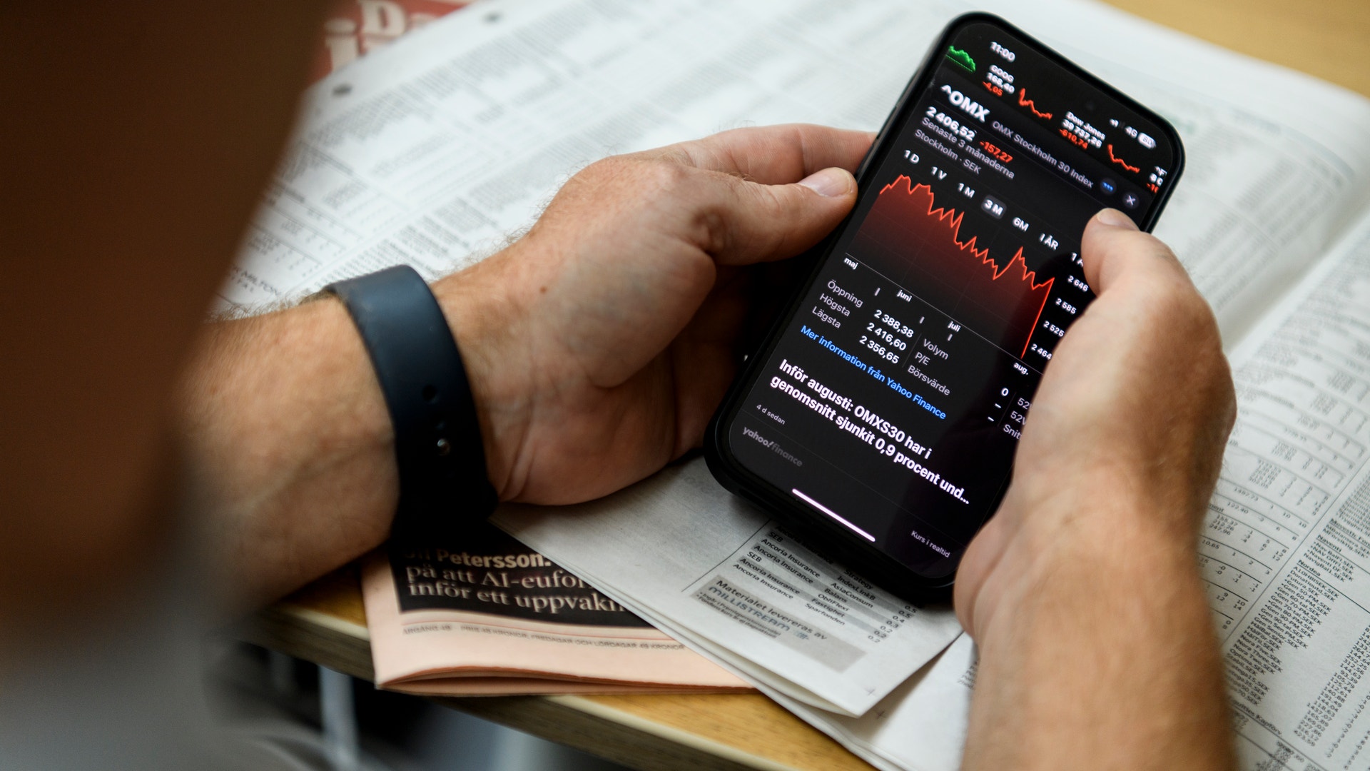 Person håller en smartphone med aktiemarknadsinformation och grafer, sittande vid ett skrivbord med finansiella dokument omkring sig.