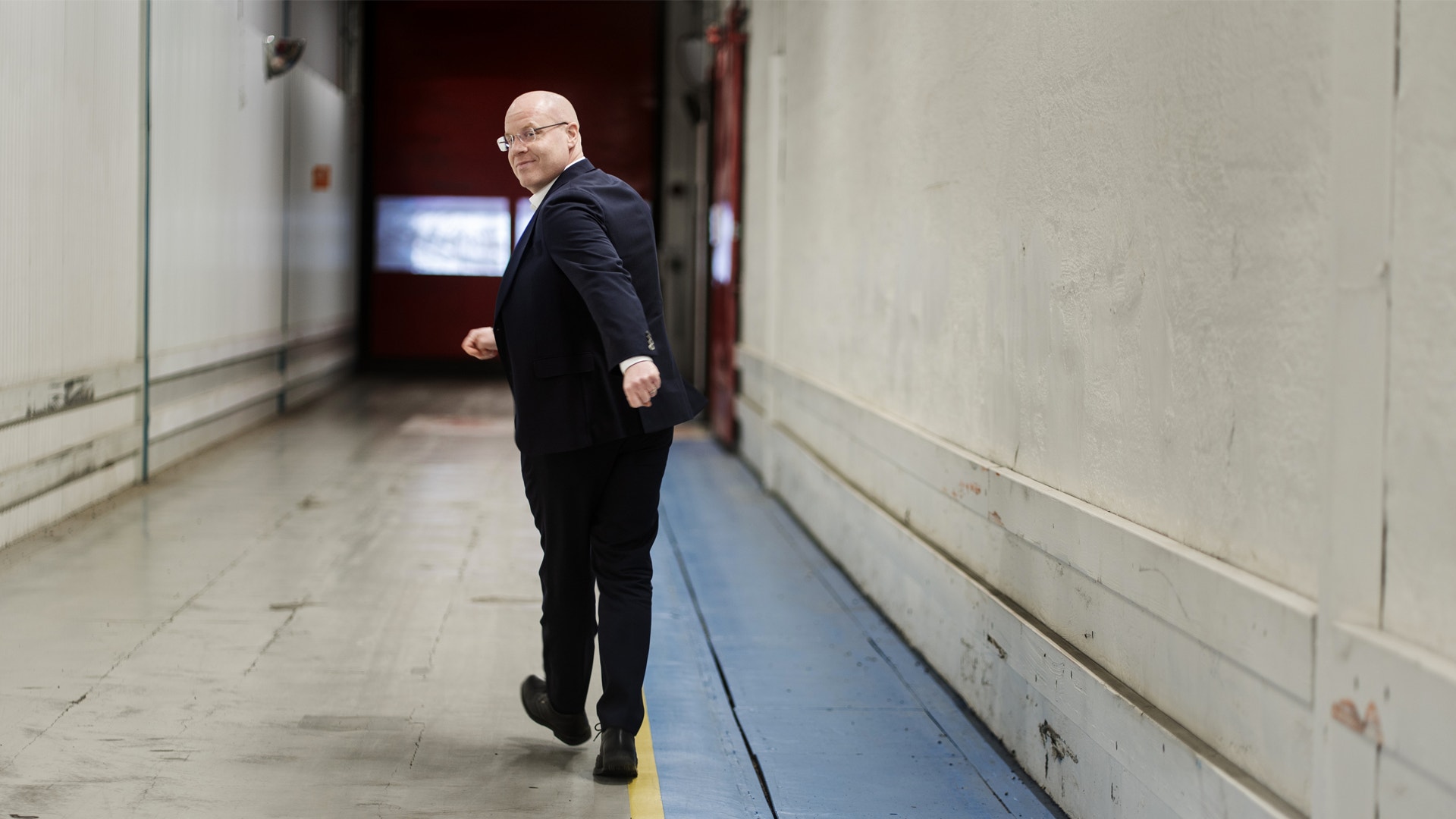 En man i mörkblå kostym och glasögon går genom en industriell korridor med vita väggar och en blå linje på golvet. Han ser över axeln medan han går in mot en dörr i bakgrunden.