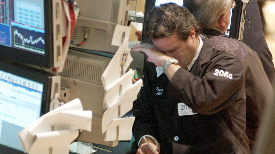 En handlare på Wall Street arbetar vid sin arbetsstation omgiven av flera datorskärmar som visar aktiekurser och marknadsdata. Handlaren är klädd i en brun jacka med "SORA" märkning och är fokuserad på att övervaka finansmarknaden. I bakgrunden syns andra finansprofessionella och ytterligare handelsterminer.