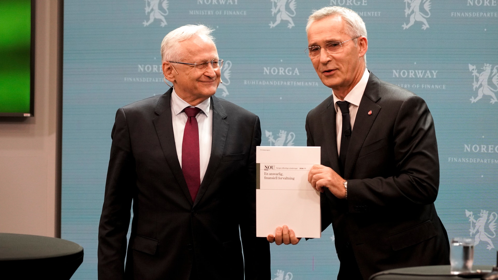 Två män i mörka kostymer står bredvid varandra framför en blå vägg med texten Norge och Finansdepartementet. Mannen till höger håller upp en rapport.