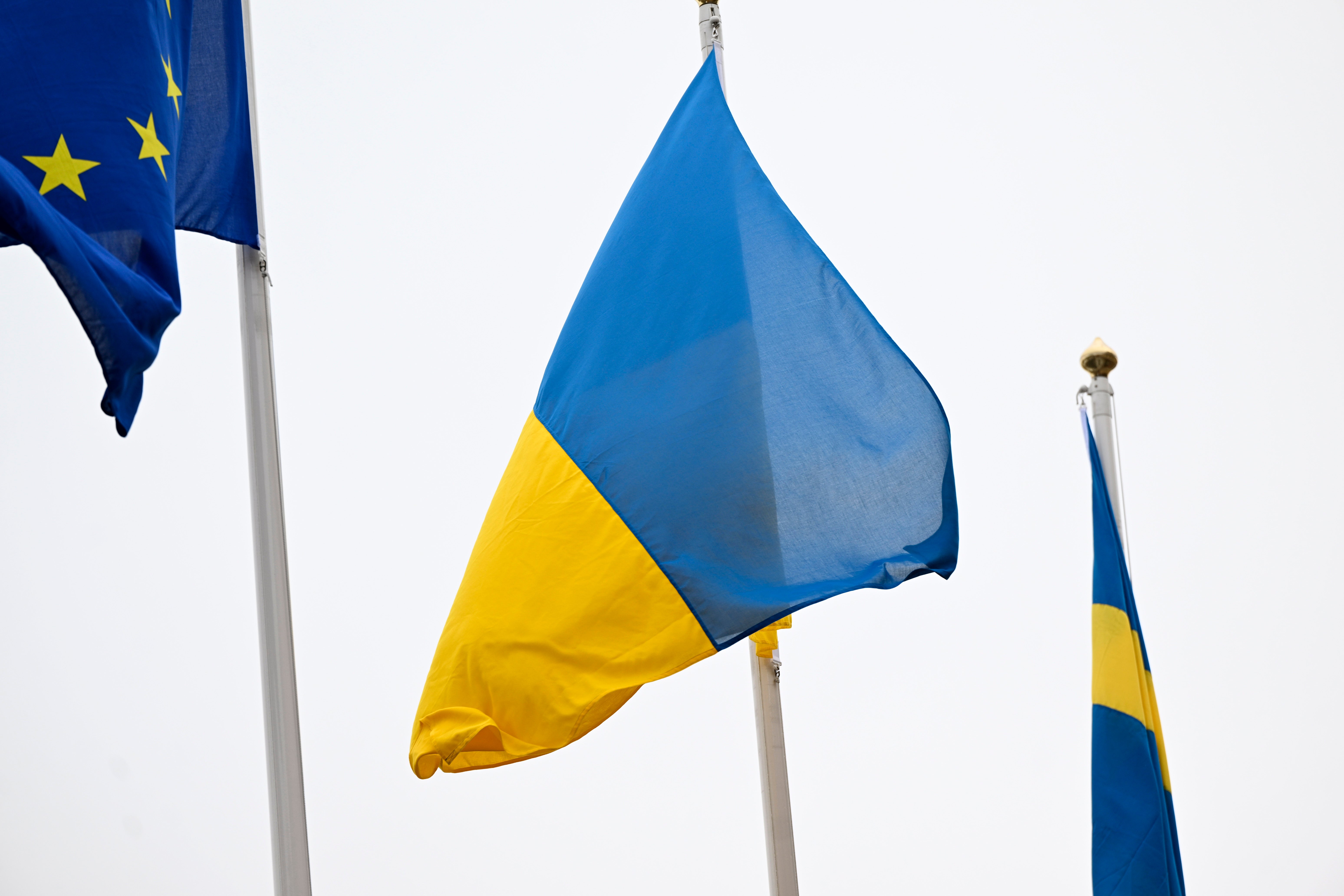 Tre flaggor som vajar i vinden: Ukrainas blå och gula flagga i mitten, EU-flaggan till vänster och en blå-gul flagga till höger.