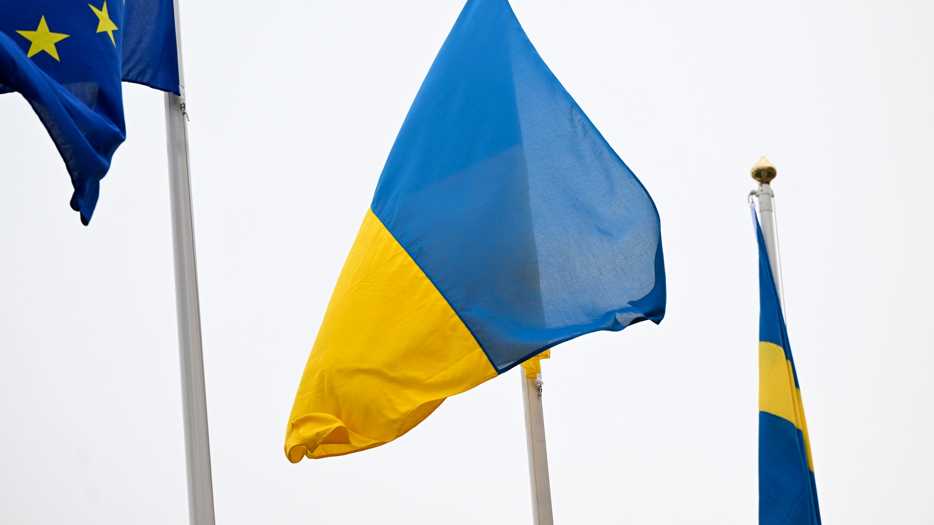 Tre flaggor som vajar i vinden: Ukrainas blå och gula flagga i mitten, EU-flaggan till vänster och en blå-gul flagga till höger.