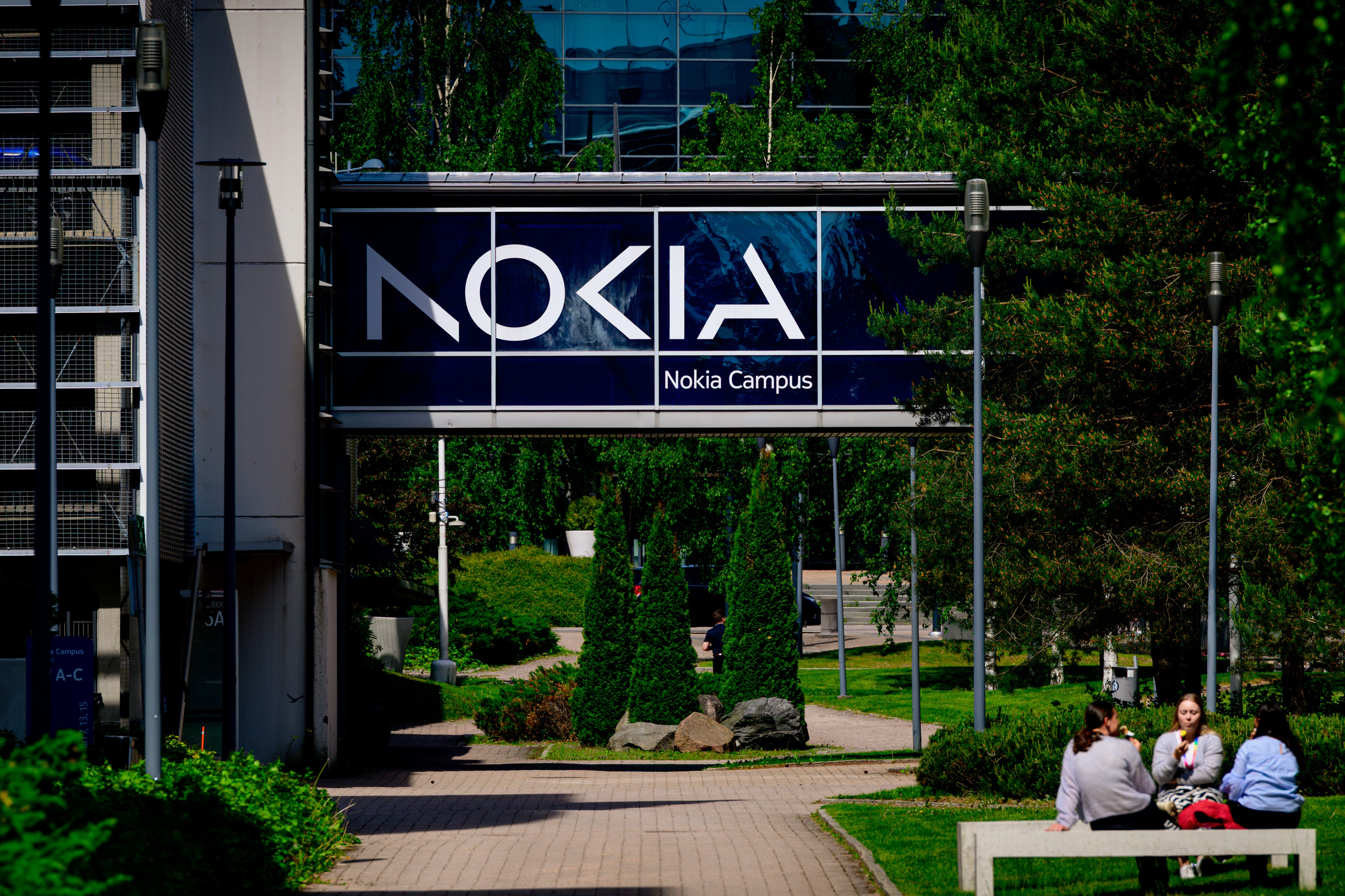 Nokia Campus-skylt med logotypen Nokia på en byggnad omgiven av gröna träd och växter. I förgrunden sitter några personer på en bänk.