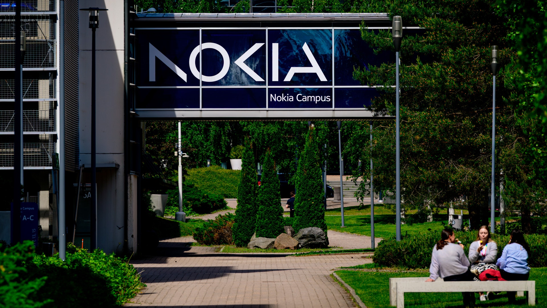 Nokia Campus-skylt med logotypen Nokia på en byggnad omgiven av gröna träd och växter. I förgrunden sitter några personer på en bänk.