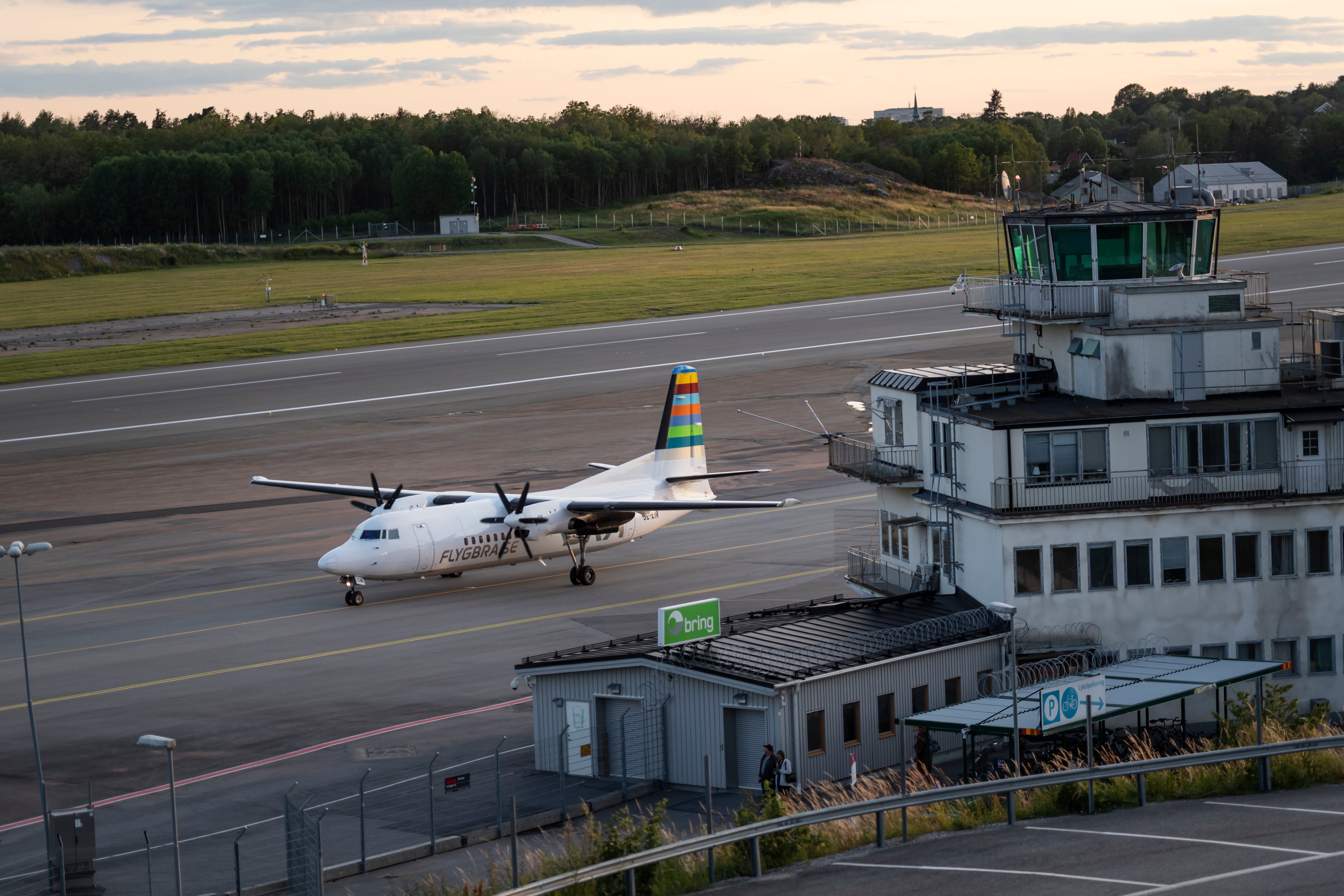 Ett turbopropdrivet passagerarflygplan står på landningsbanan vid en flygplats. I förgrunden syns en kontrolltorn och flygplatsbyggnader. I bakgrunden ses gröna ängar och barrskog.