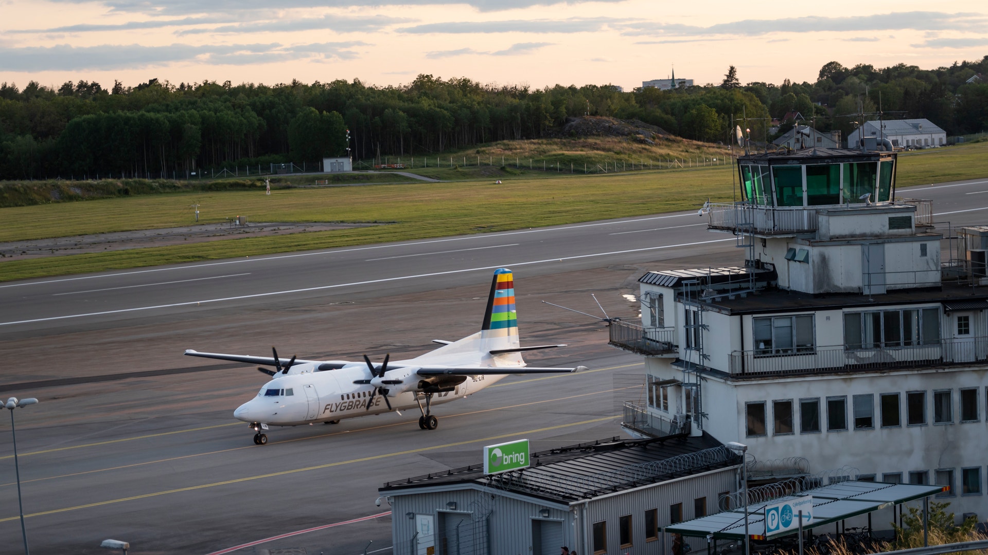 Ett turbopropdrivet passagerarflygplan står på landningsbanan vid en flygplats. I förgrunden syns en kontrolltorn och flygplatsbyggnader. I bakgrunden ses gröna ängar och barrskog.