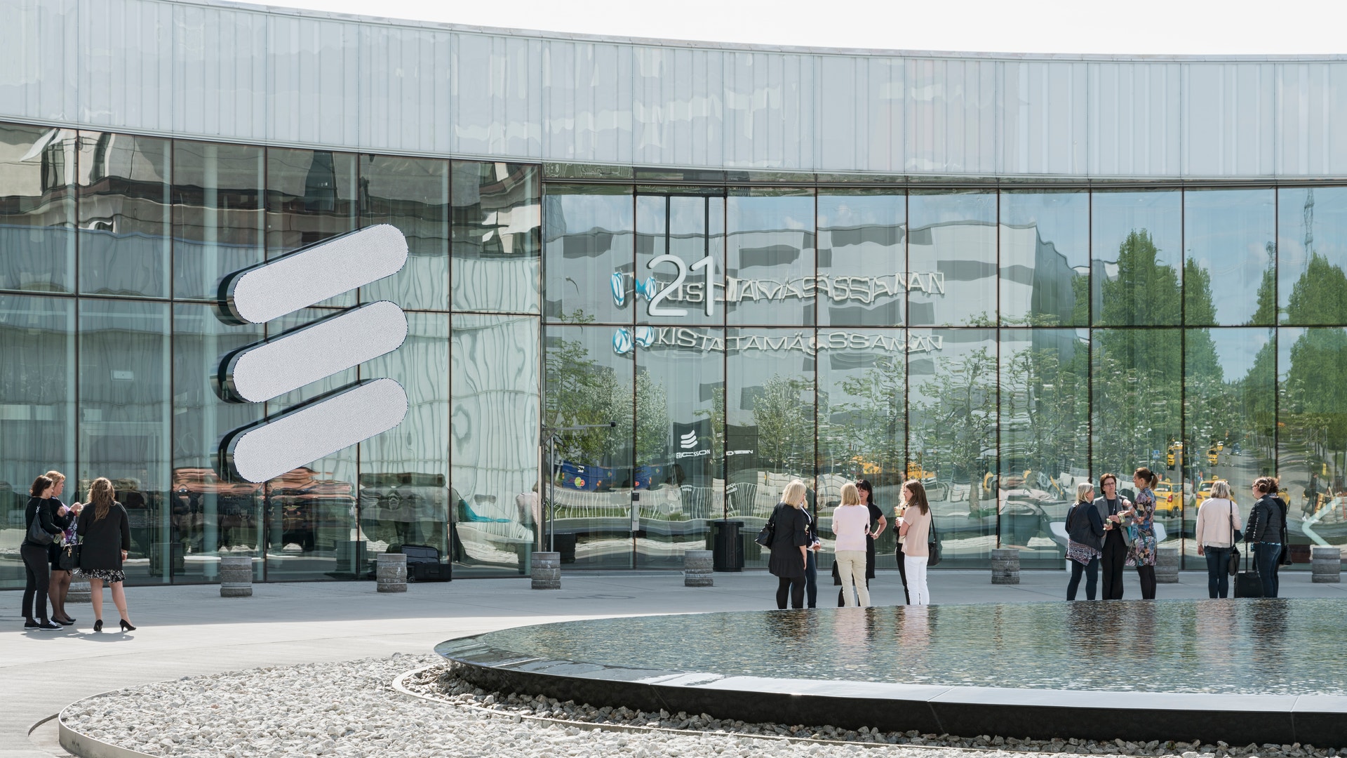 Ericssons huvudkontor med modern glasfasad och vita logotypstreck. Framför byggnaden finns en öppen plats med vattenspegel och människor.