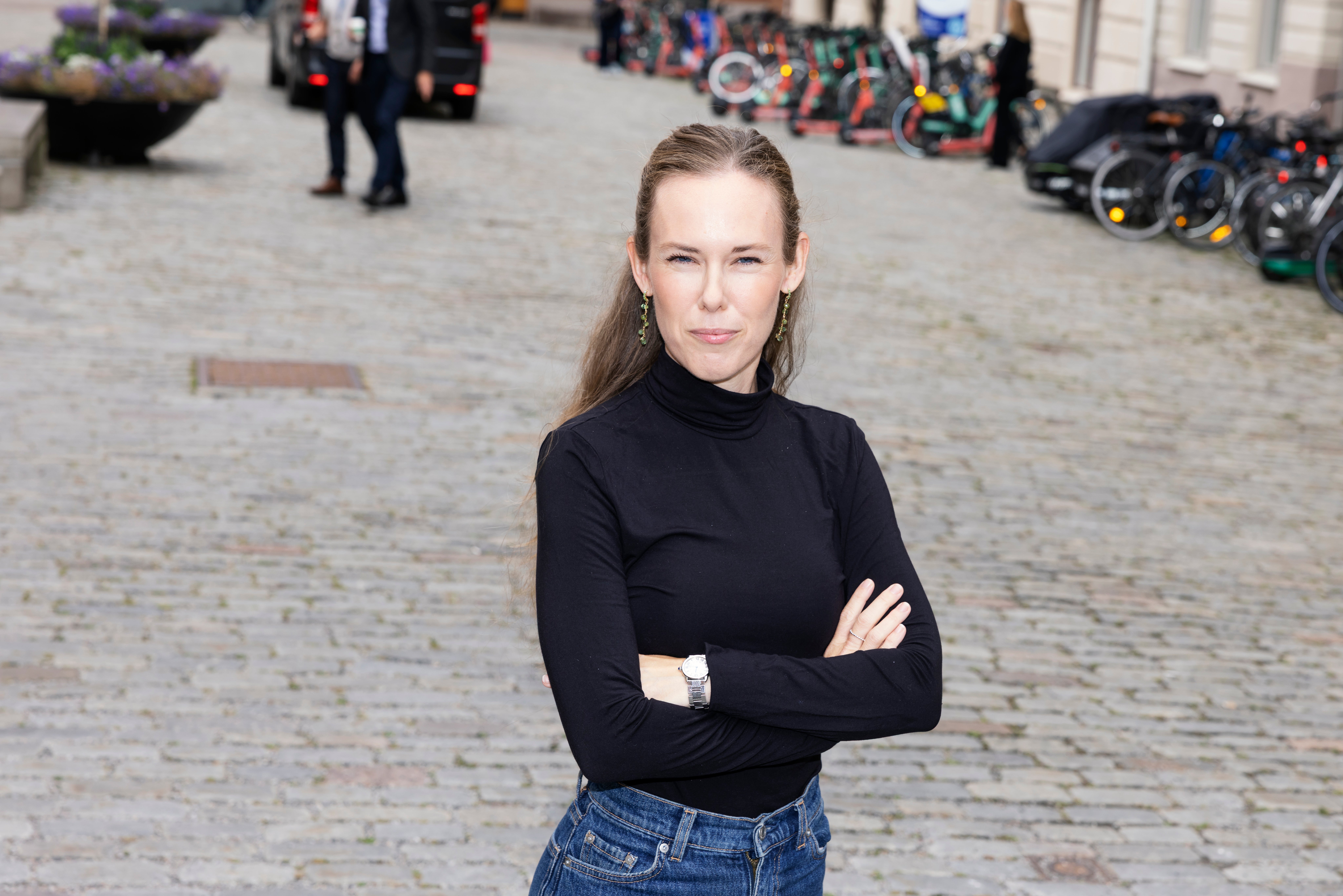 Porträtt av en kvinna som står på en kullerstenbelagd gata. Hon är klädd i svart polotröja och jeans, med armarna i kors.