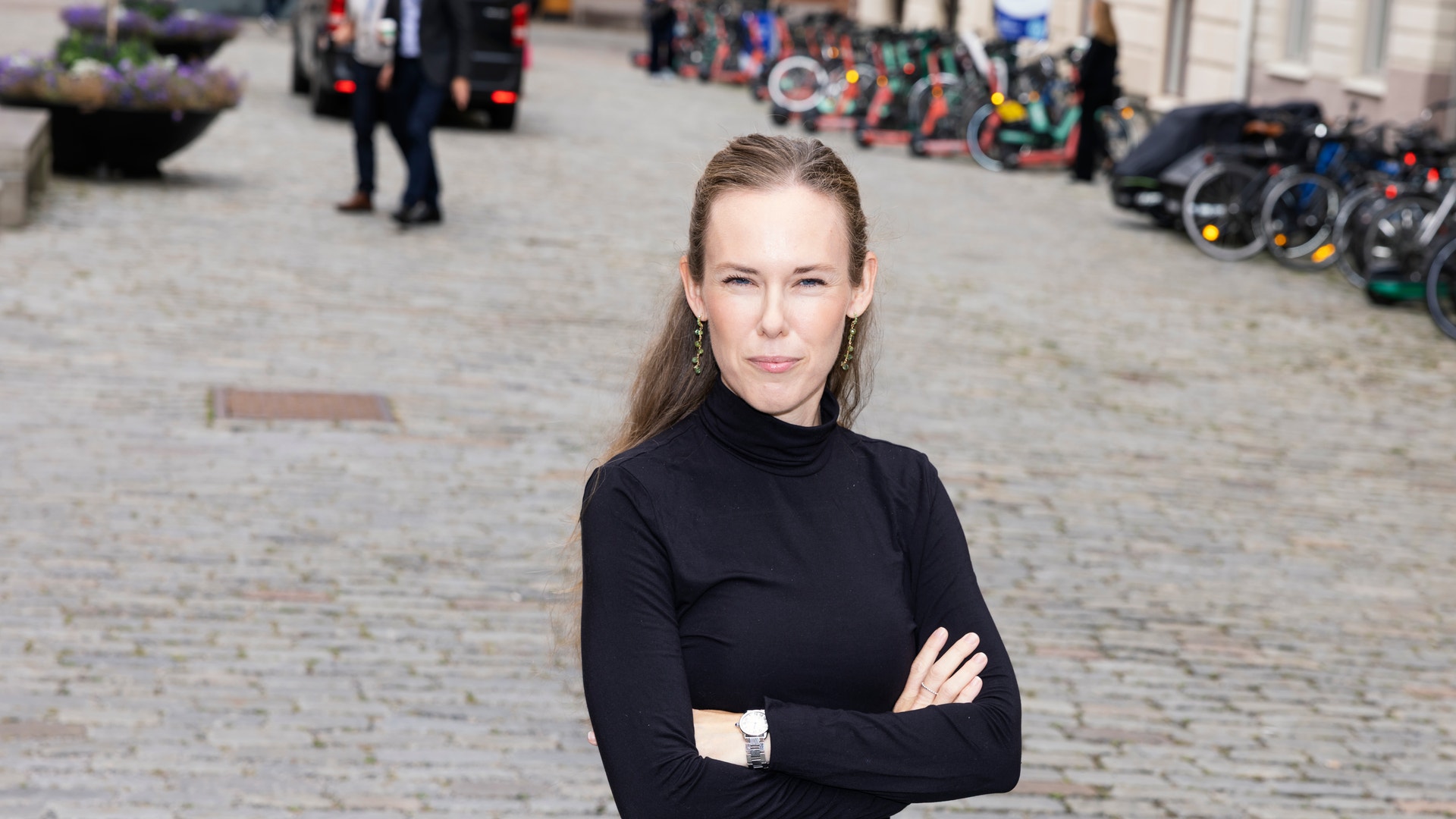 Porträtt av en kvinna som står på en kullerstenbelagd gata. Hon är klädd i svart polotröja och jeans, med armarna i kors.