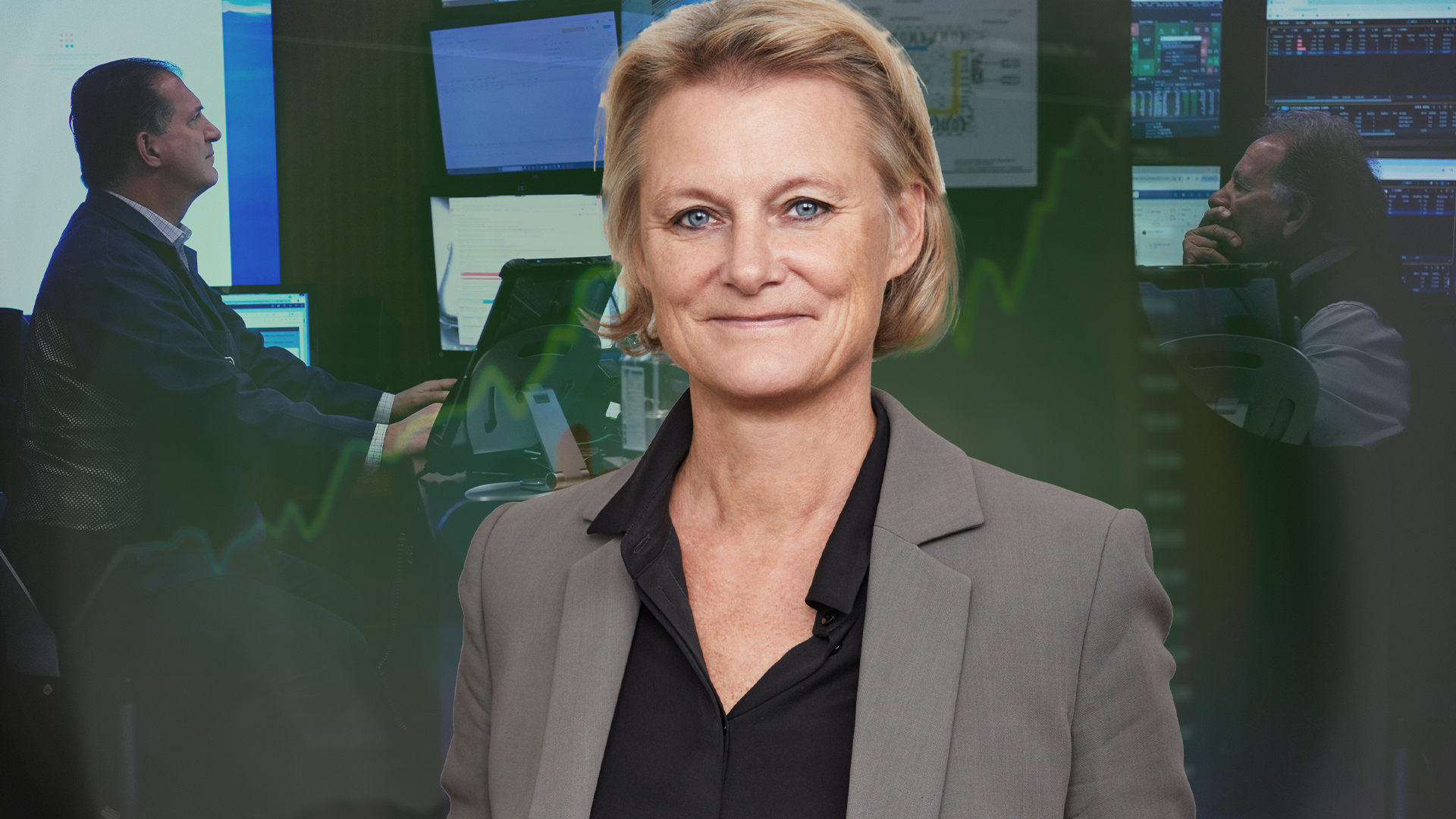 Porträttbild av en kvinna i grå blazer som ler mot kameran. I bakgrunden arbetar andra personer vid datorer i ett modernt kontorsrum med gröna väggar.