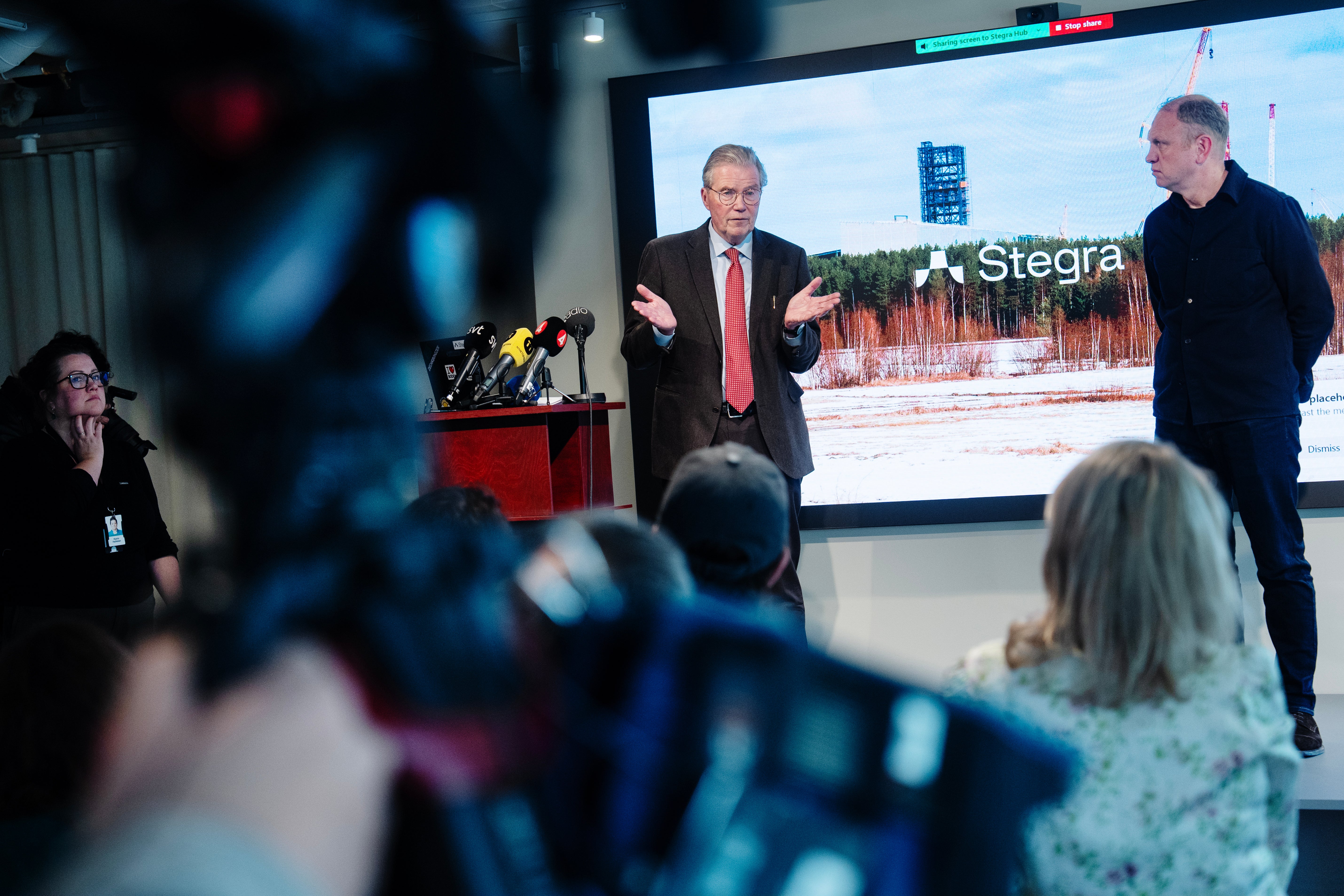 En man i kostym och röd slips presenterar vid ett podium under en presskonferens, medan en annan man står vid hans sida. En skärm bakom dem visar en presentation med Stegra-logotypen. Publiken sitter framför scenen och lyssnar.
