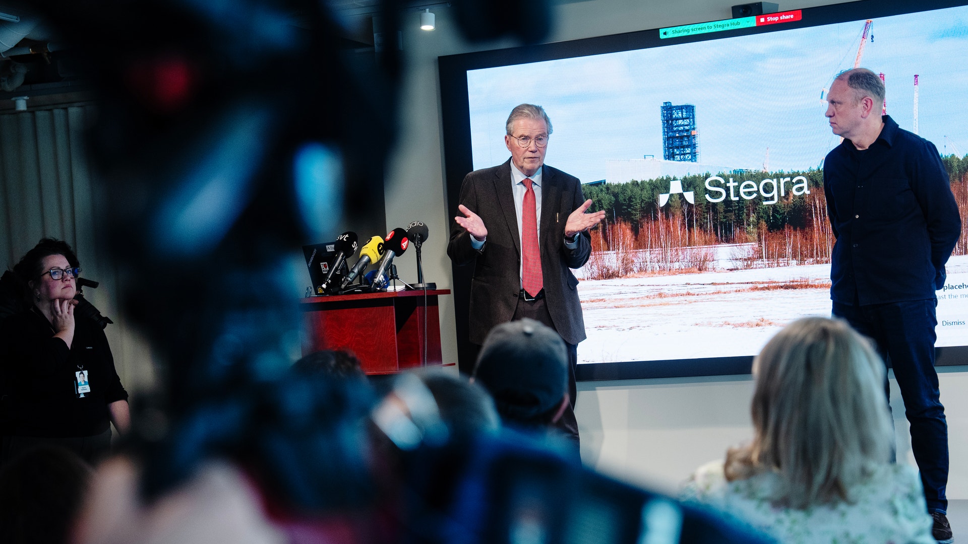 En man i kostym och röd slips presenterar vid ett podium under en presskonferens, medan en annan man står vid hans sida. En skärm bakom dem visar en presentation med Stegra-logotypen. Publiken sitter framför scenen och lyssnar.
