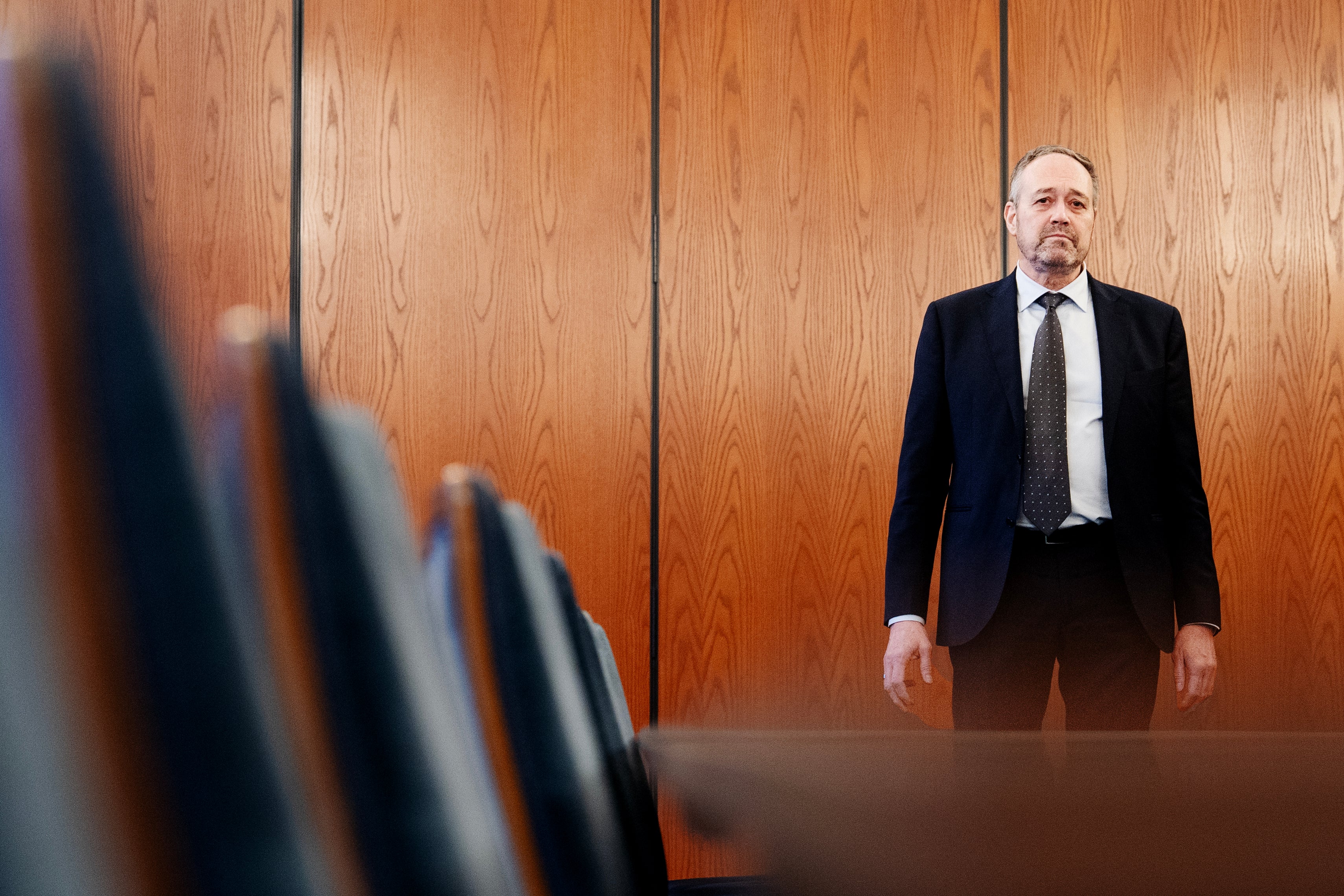 Man i kostym står framför en trävägg med suddig publik i förgrunden.