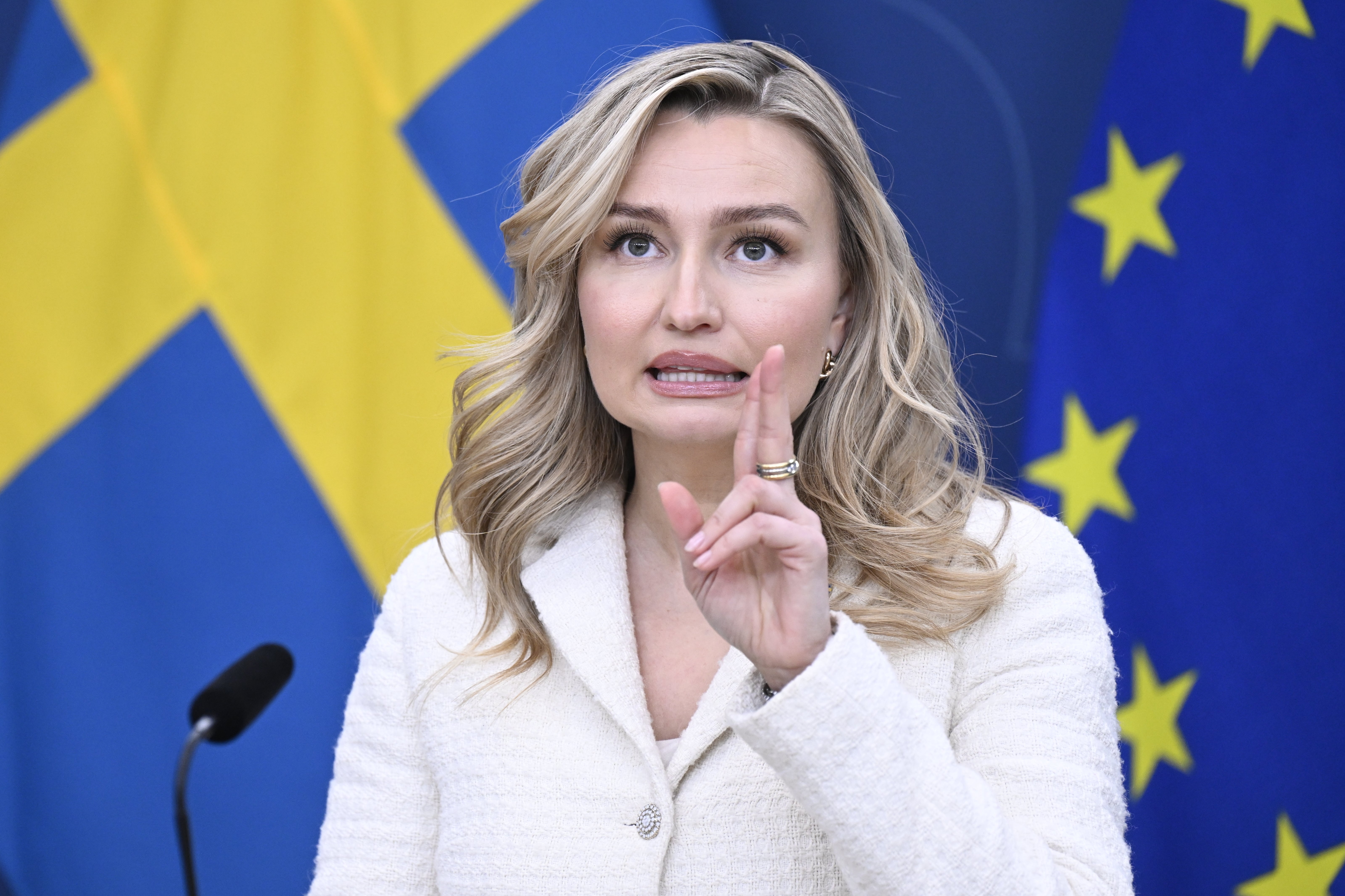 Kvinnlig person i vit kavaj framför bakgrund med svenska flaggan och EU-flaggan.