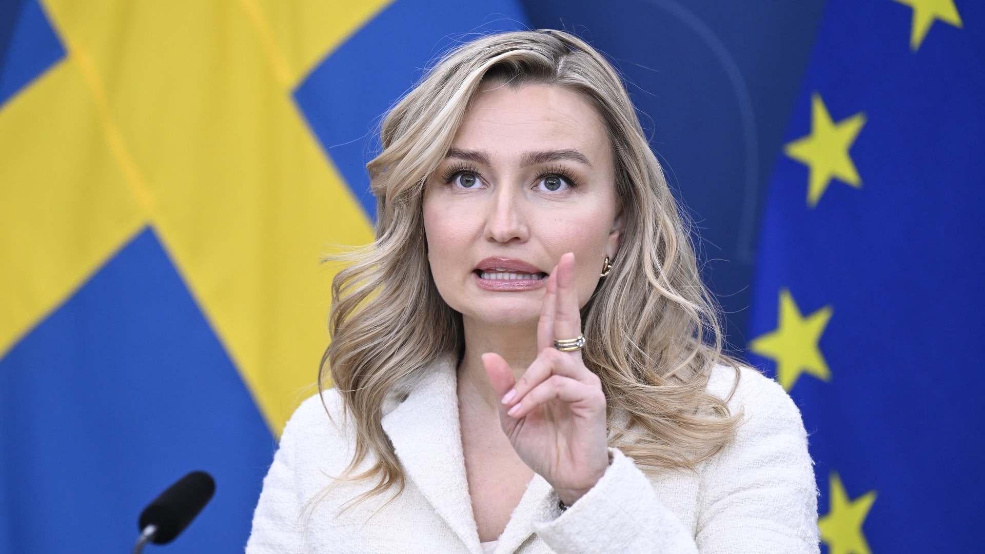 Kvinnlig person i vit kavaj framför bakgrund med svenska flaggan och EU-flaggan.