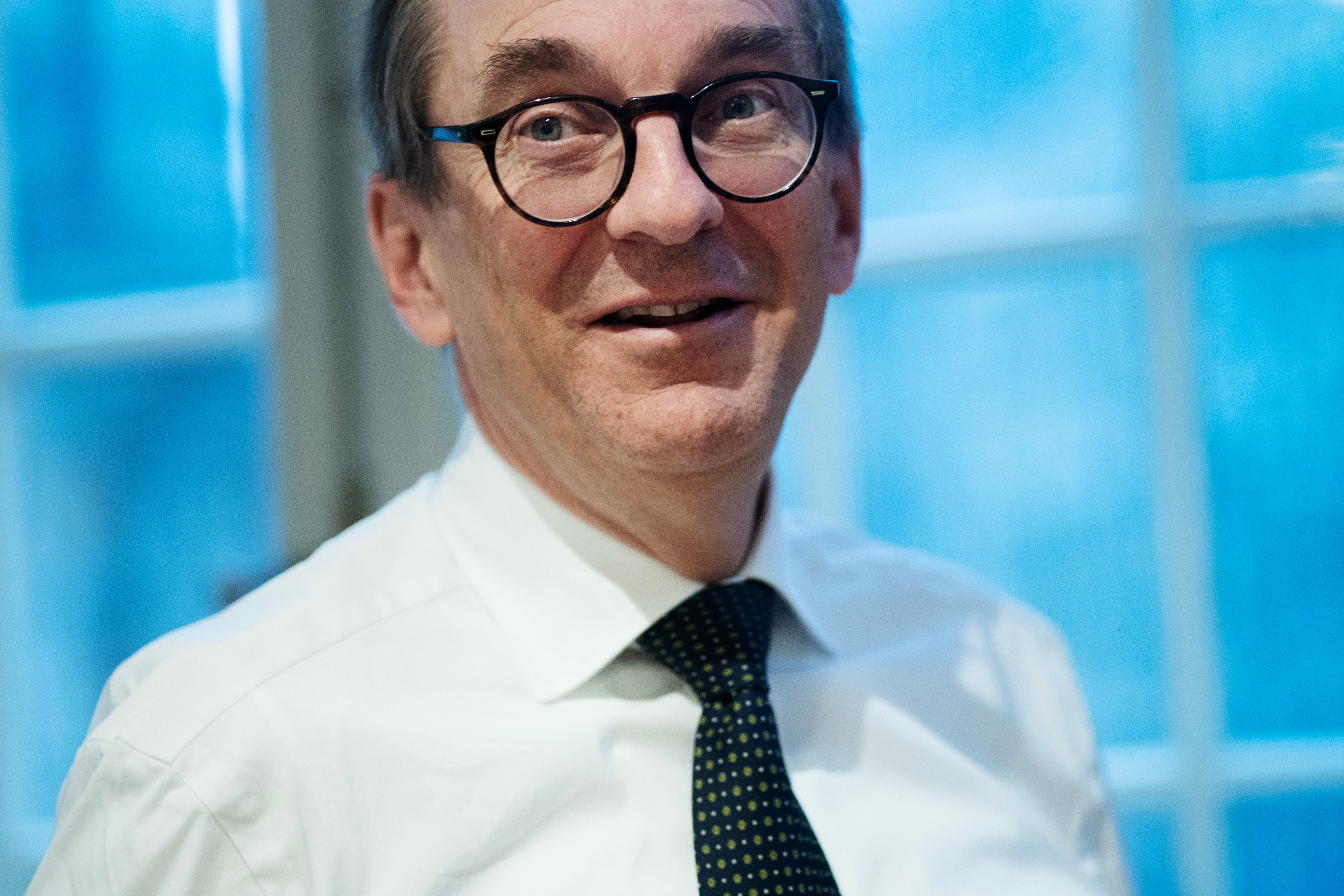 Porträttfoto av en man med glasögon, vit skjorta och mörkblå slips framför ett blått fönster.