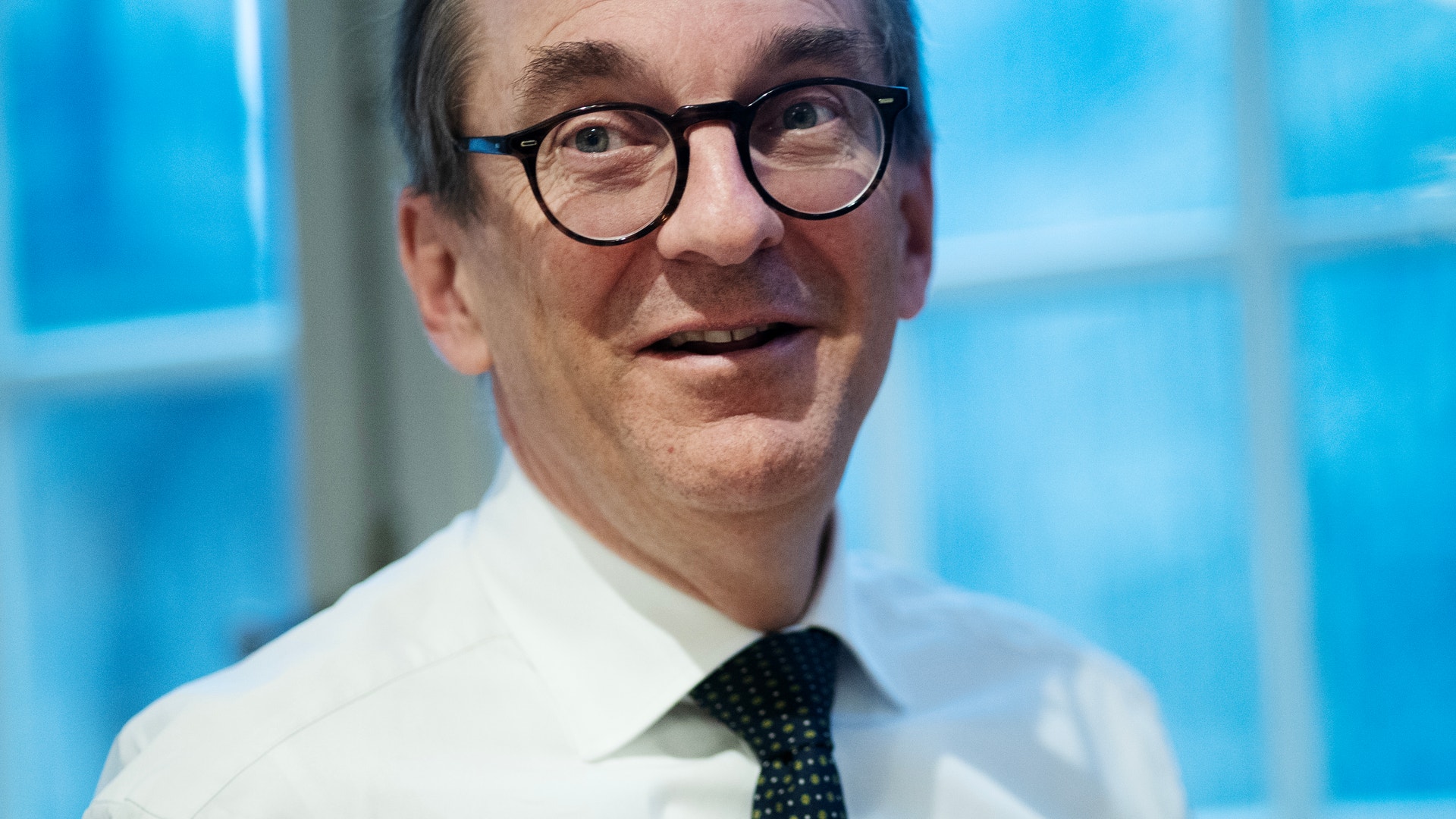 Porträttfoto av en man med glasögon, vit skjorta och mörkblå slips framför ett blått fönster.
