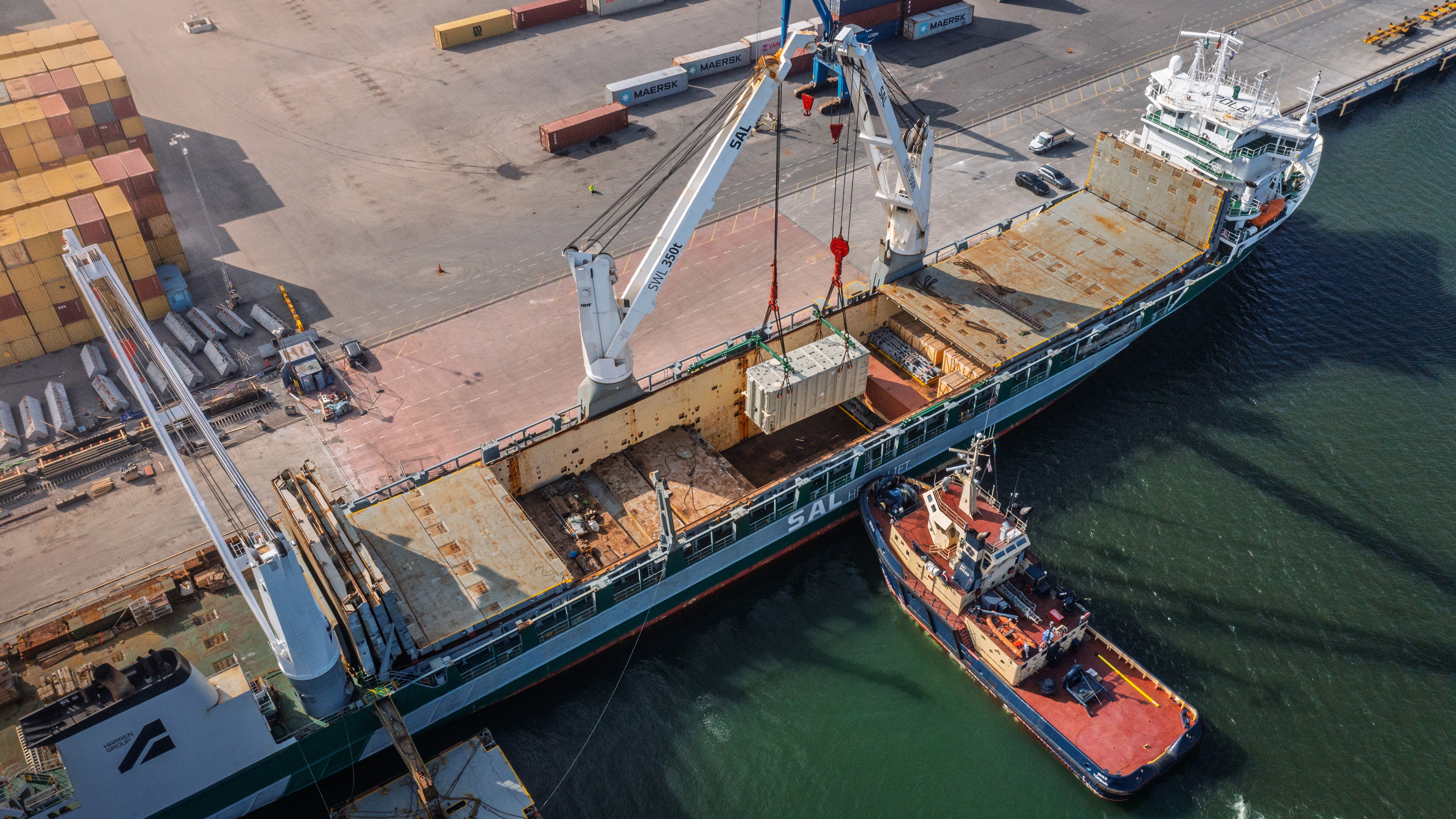 Flygfoto av en hamn där en stor lastfartyg lastas med hjälp av en kran. En röd bogserbåt ligger bredvid fartyget. Containrar och hamnutrustning syns på kajen.