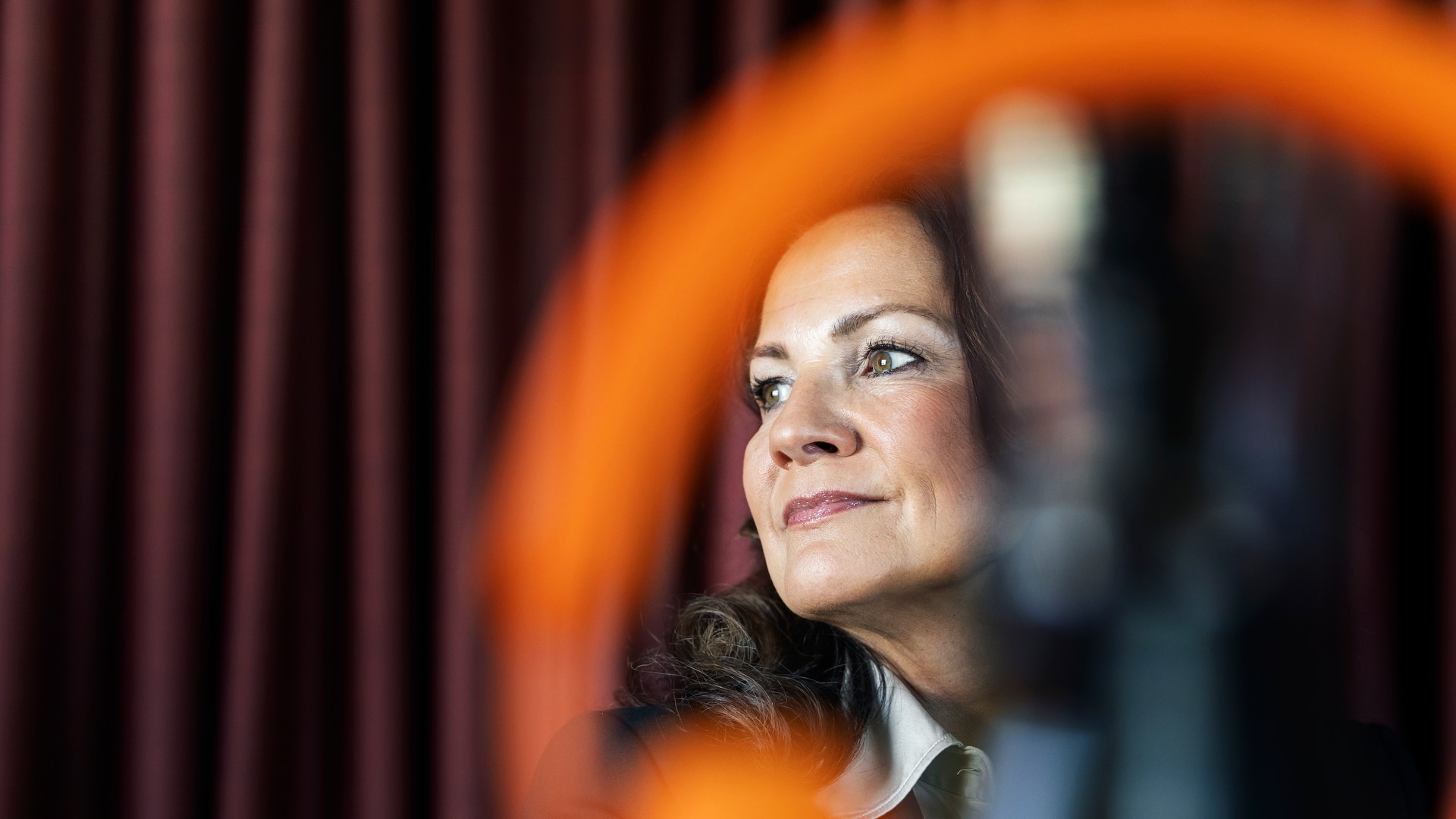Porträtt av en kvinna som ser ut mot sidan framför en röd gardin. En orange cirkulär form är suddig i förgrunden.