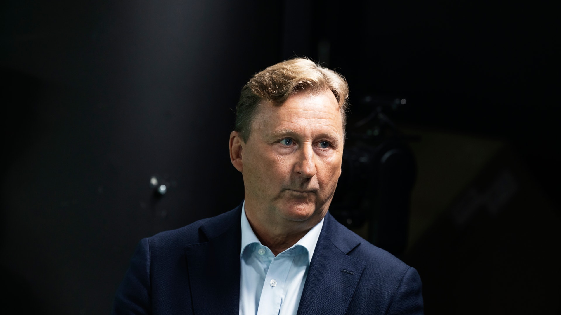 Porträtt av en man i mörk kostym och ljus skjorta mot mörk bakgrund.