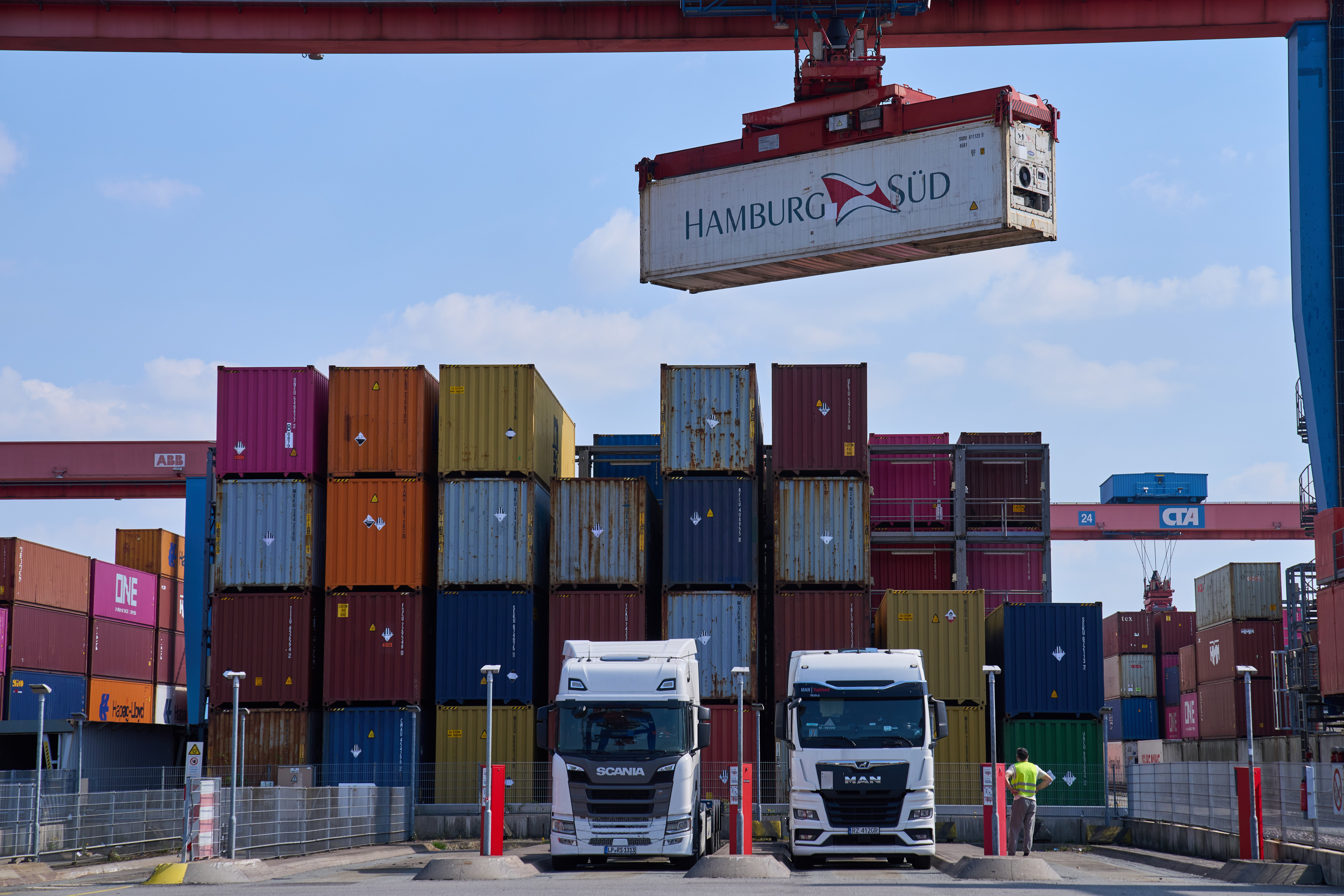 Två lastbilar står framför en stor hög med färgglada containerfartygscontainrar på en containerhamn. En röd kran och en skylt med texten HAMBURG SÜD syns ovanför containrarna.