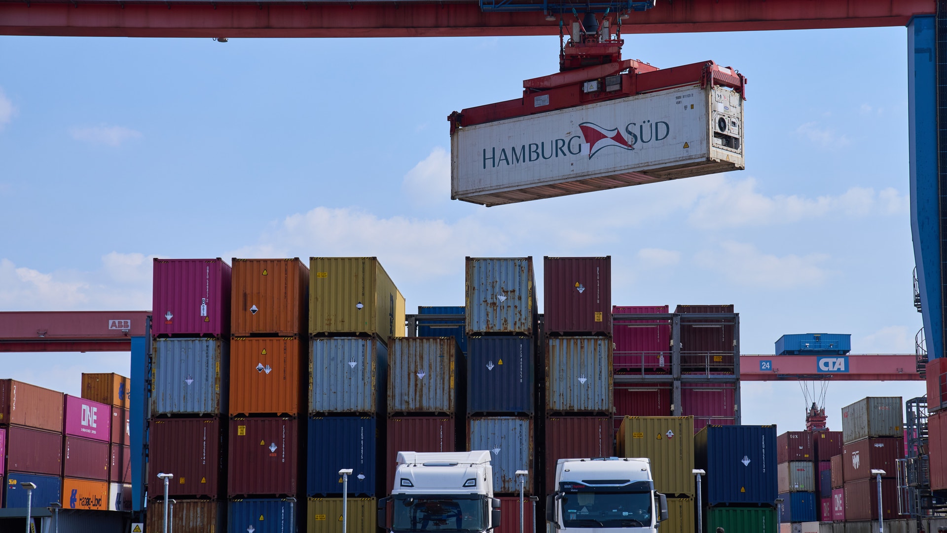 Två lastbilar står framför en stor hög med färgglada containerfartygscontainrar på en containerhamn. En röd kran och en skylt med texten HAMBURG SÜD syns ovanför containrarna.