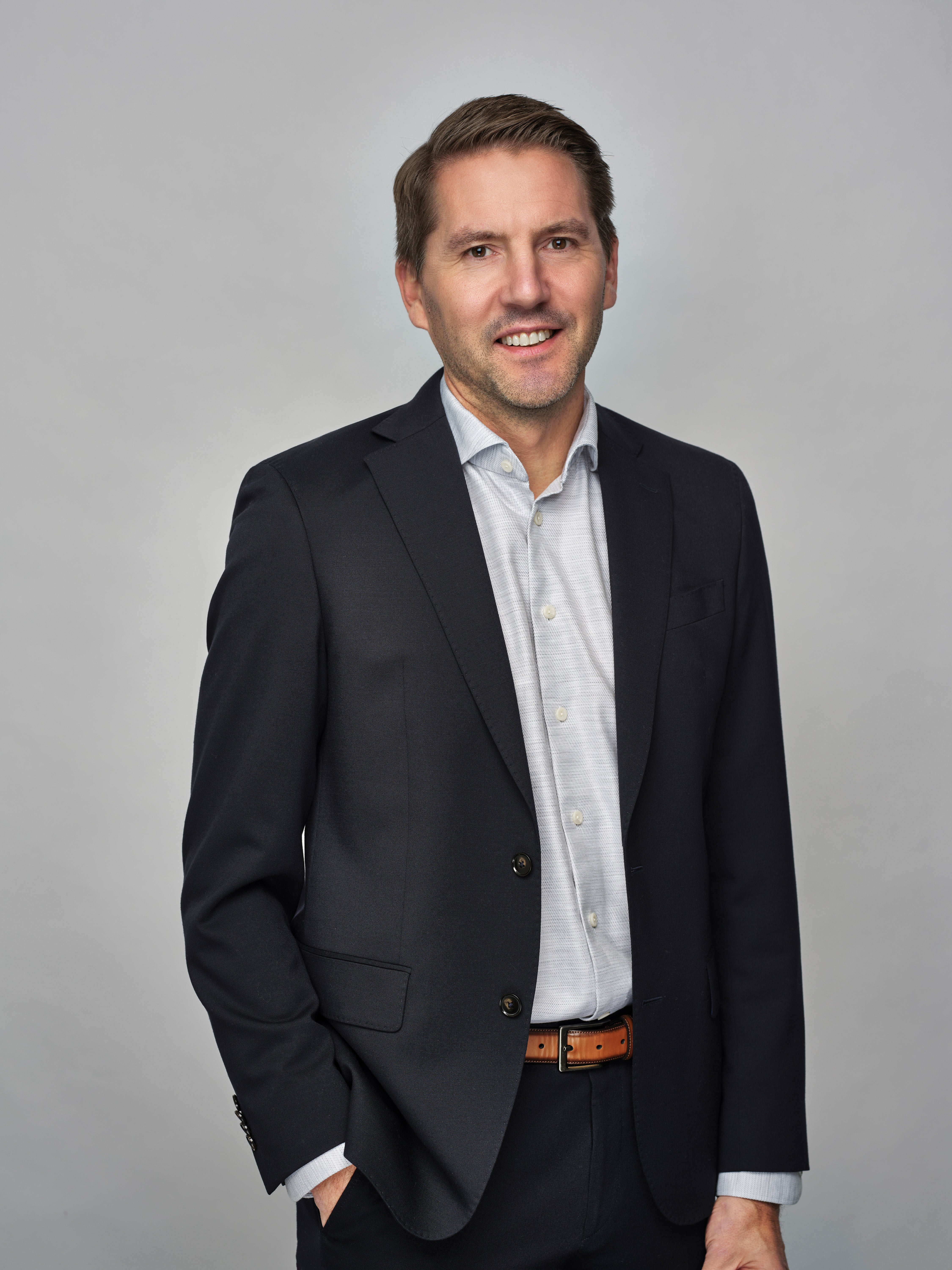 Porträtt av Johan Andersson, President and CEO. Han bär en svart kavaj och ljusblå skjorta med orange bälte, och ler mot kameran mot en grå bakgrund.