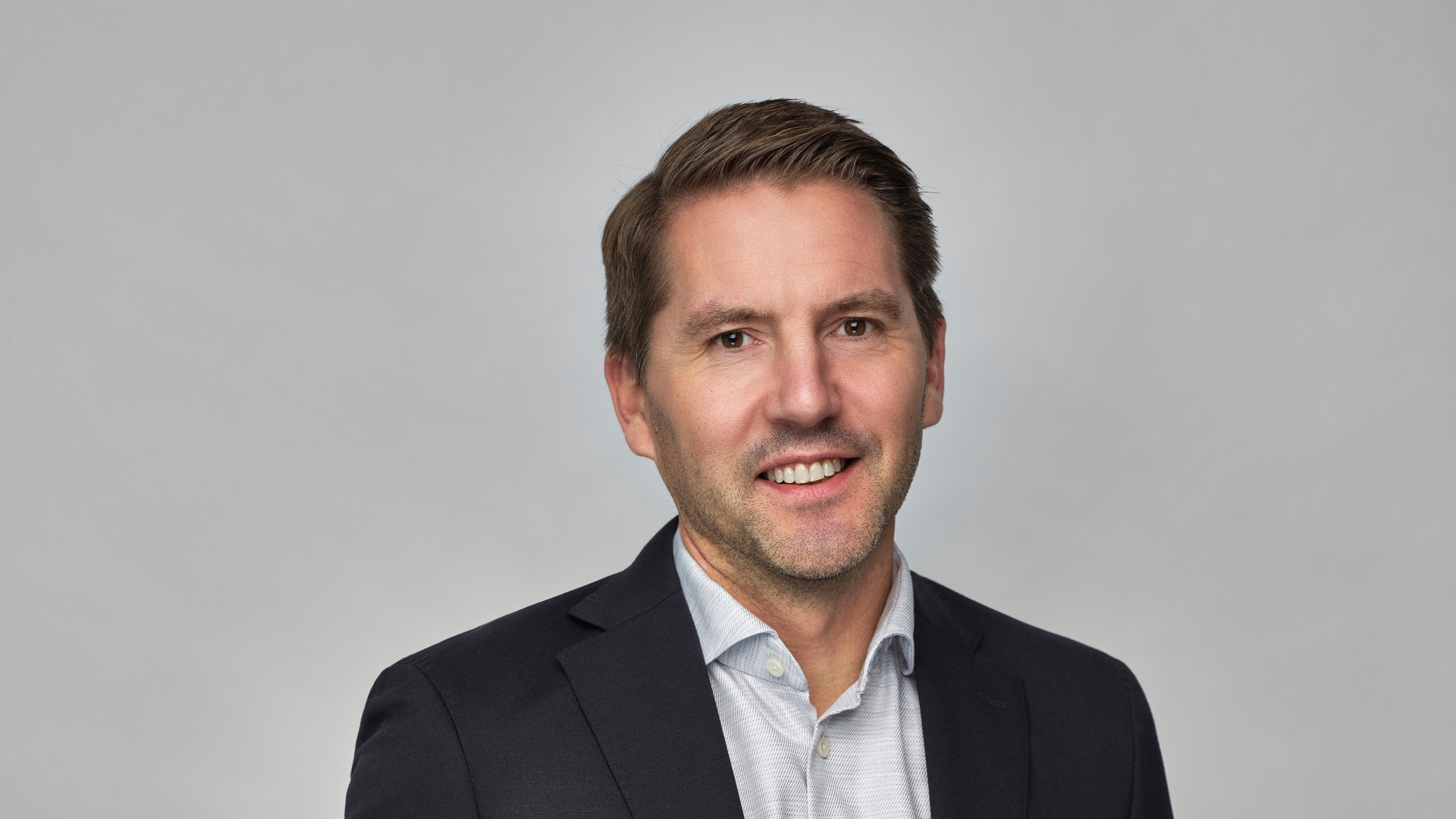 Porträtt av Johan Andersson, President and CEO. Han bär en svart kavaj och ljusblå skjorta med orange bälte, och ler mot kameran mot en grå bakgrund.