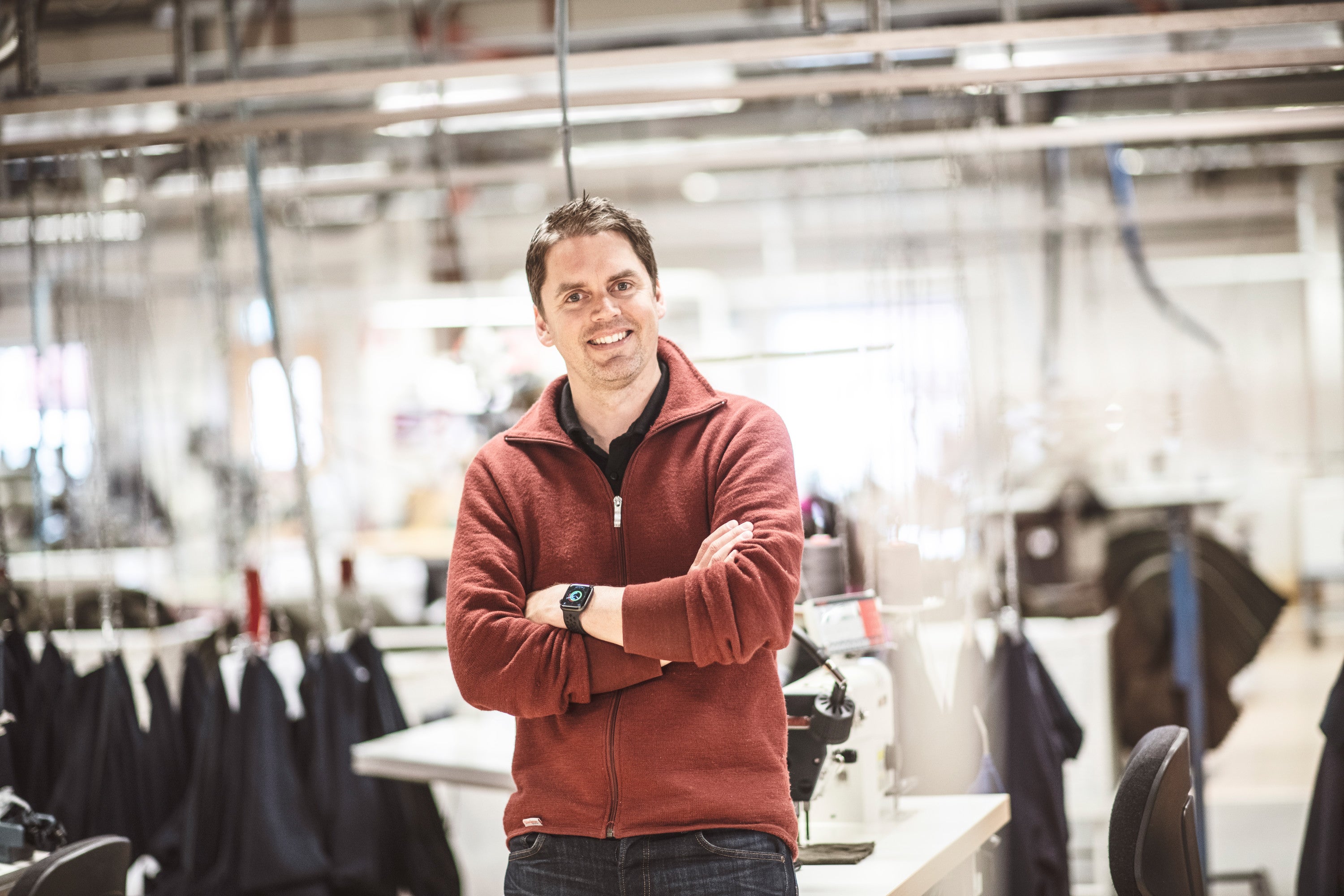 En man i rödbrun tröja står med armarna i kors och ler i en textilfabrik med symaskiner och kläder i bakgrunden.