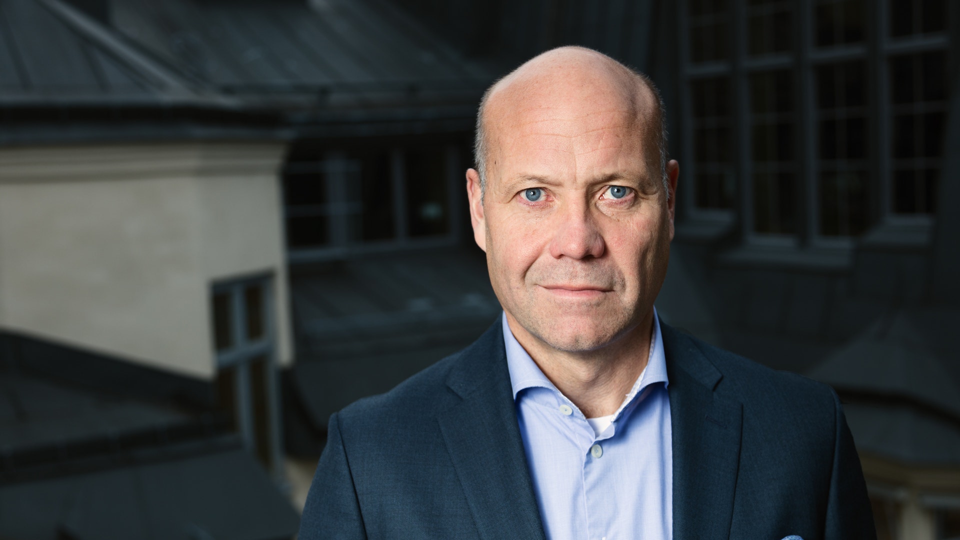 Porträtt av en kal man i mörk kavaj och ljusblå skjorta som tittar rakt in i kameran, med takåsar i bakgrunden.