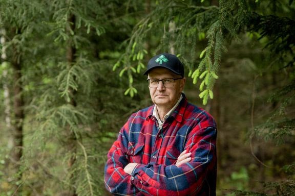 En man i rutigt skjorta och keps står framför barrträd i skogen.