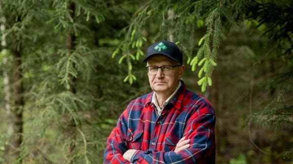 En man i rutigt skjorta och keps står framför barrträd i skogen.