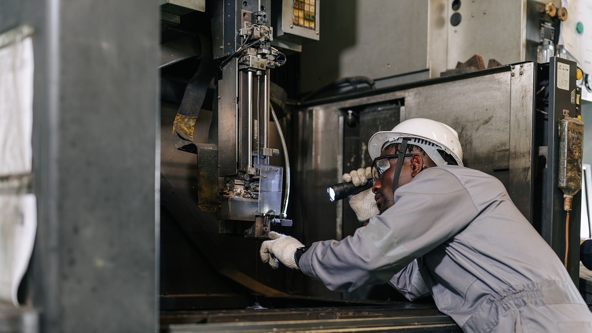 En person i skyddsutrustning inspekterar en industrimaskin med en ficklampa.