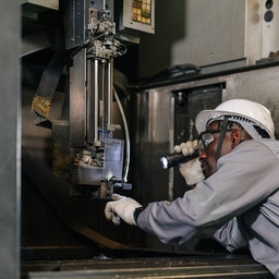 En person i skyddsutrustning inspekterar en industrimaskin med en ficklampa.