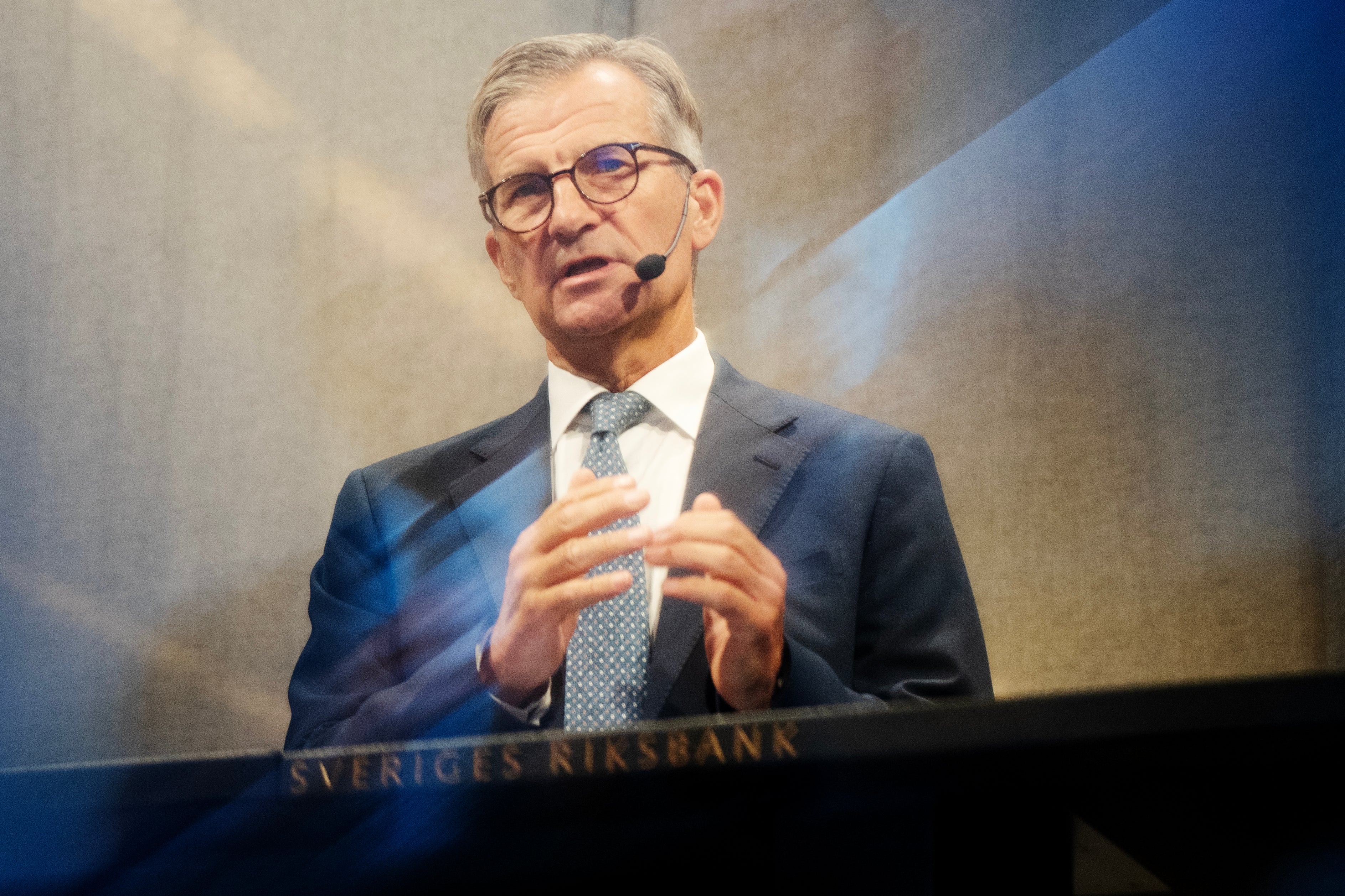 Man i kostym och glasögon som håller ett föredrag med headset-mikrofon. I bakgrunden syns texten Sveriges Riksbank.