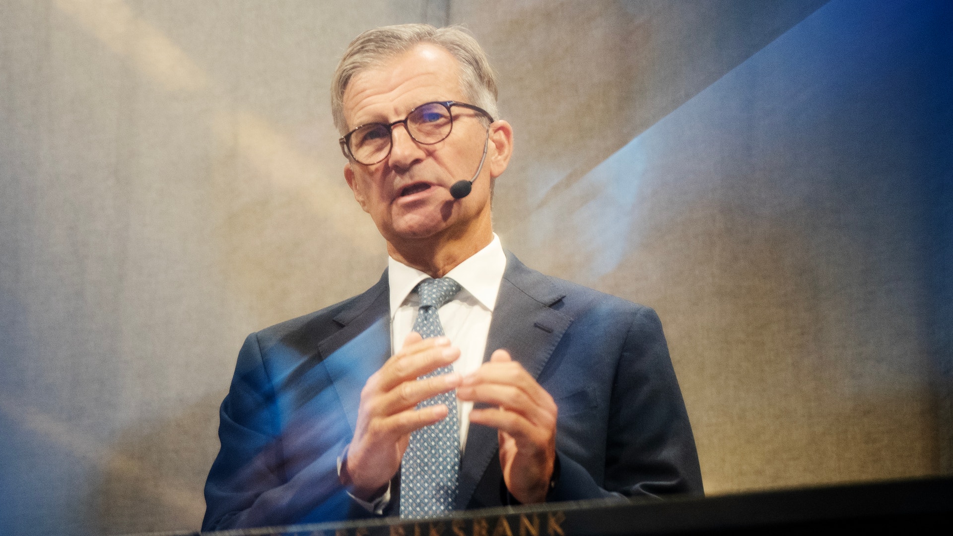 Man i kostym och glasögon som håller ett föredrag med headset-mikrofon. I bakgrunden syns texten Sveriges Riksbank.