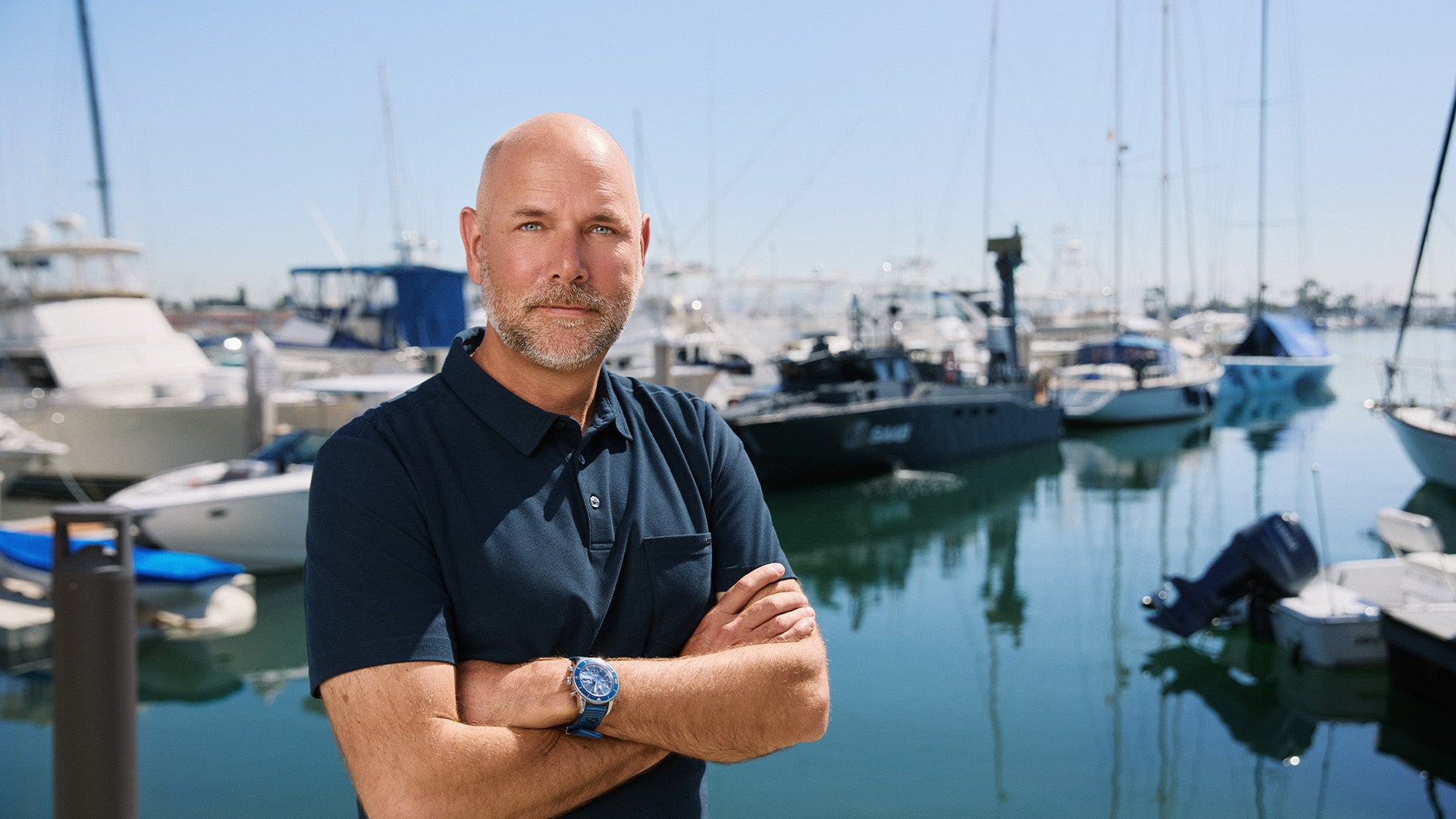Man står vid en båthamn med armarna i kors och tittar mot kameran. Flera båtar syns i bakgrunden.